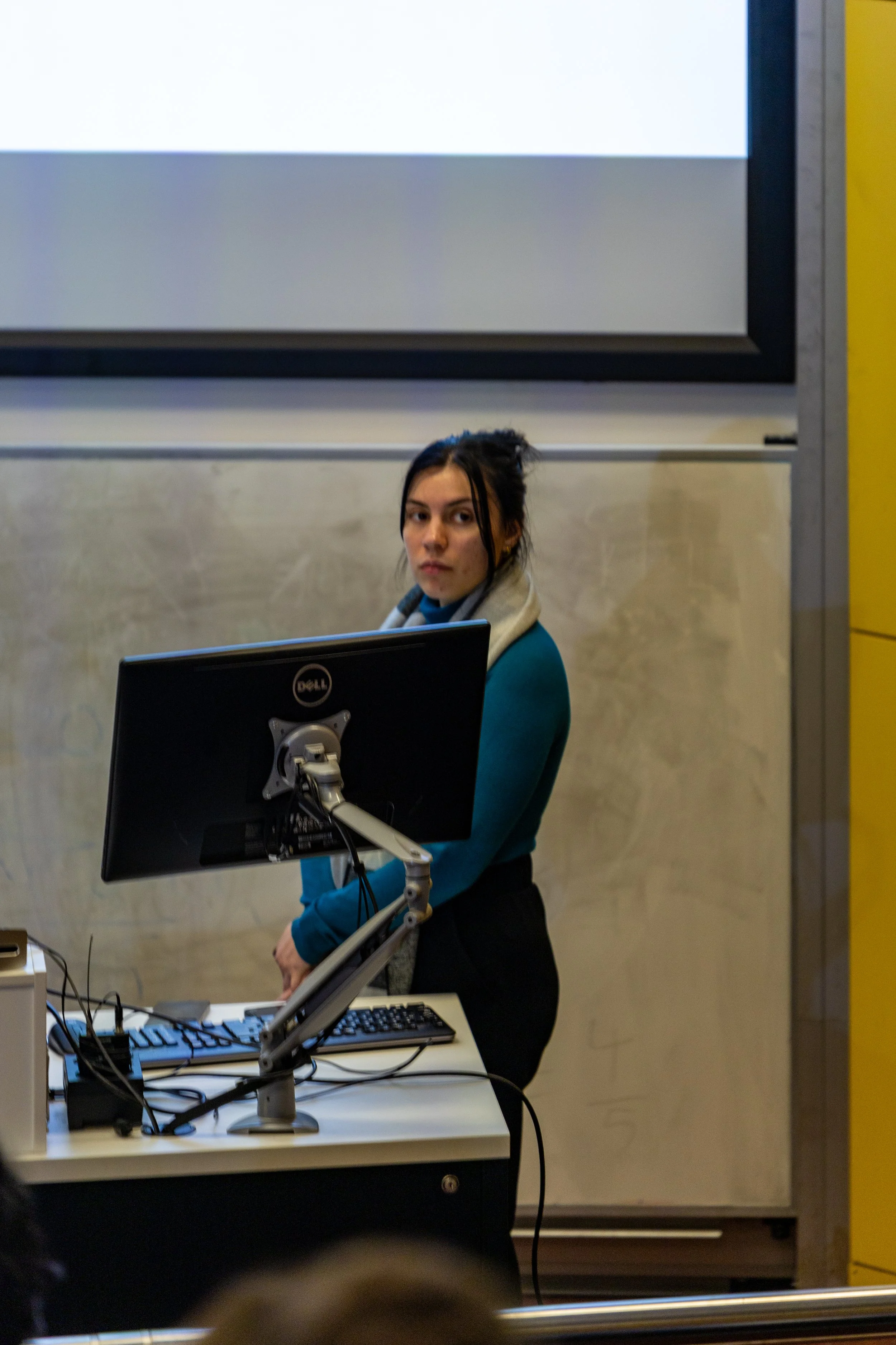 A woman standing behind a computer monitor in a classroom or conference room.