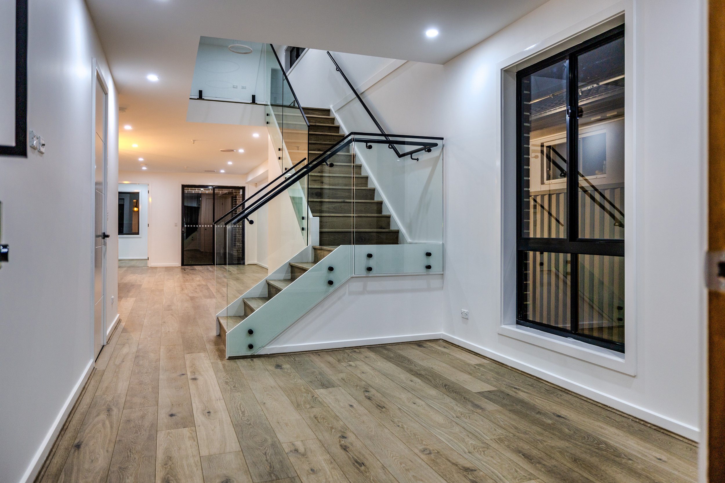 Interior of a modern house with a wooden floor, white walls, a staircase with glass railing, and large windows.