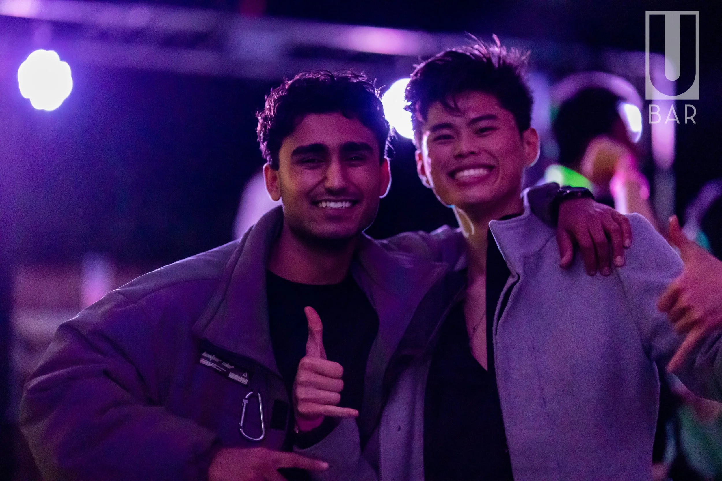 Two young men smiling and hugging each other in a dimly lit bar with purple lighting.
