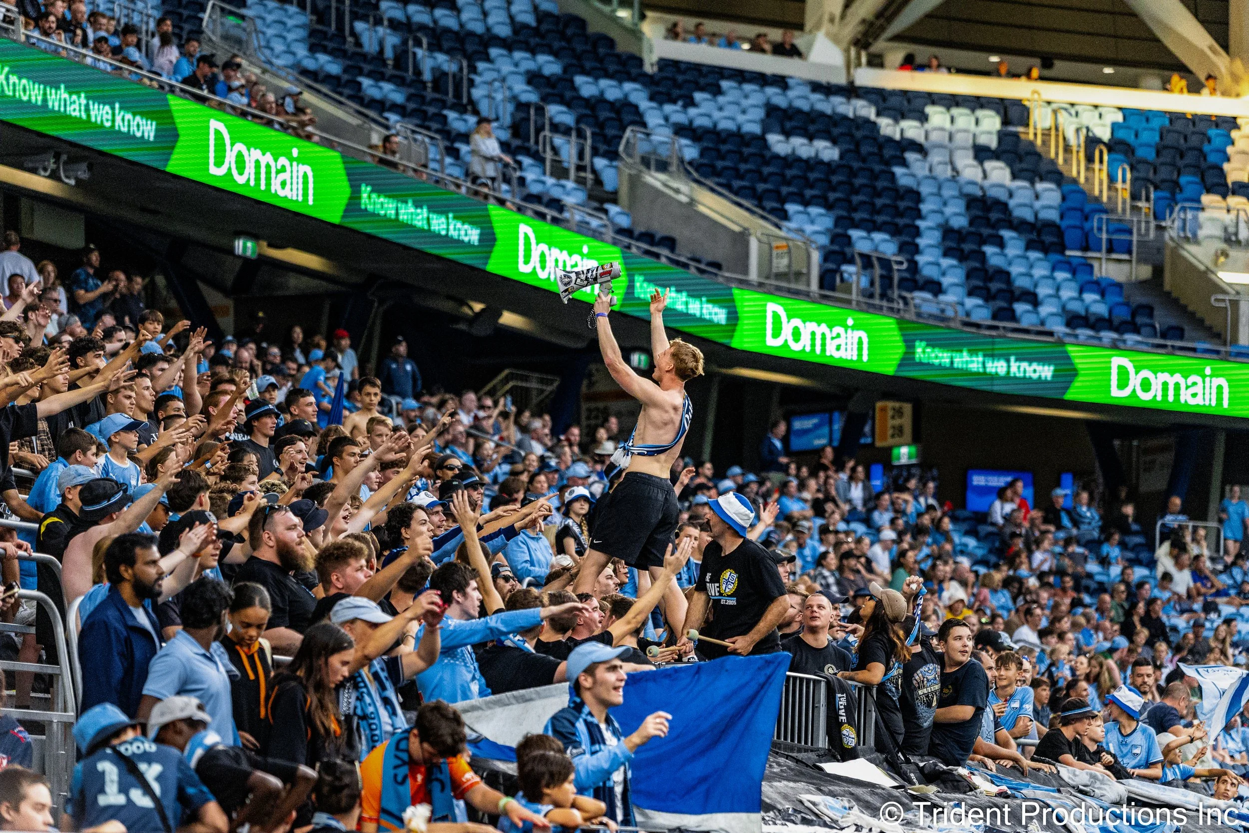 A crowd of sports fans in blue and black attire cheering as a shirtless man holds a megaphone and celebrates on a stadium bleacher. The stadium has blue seats and a green electronic advertising banner.