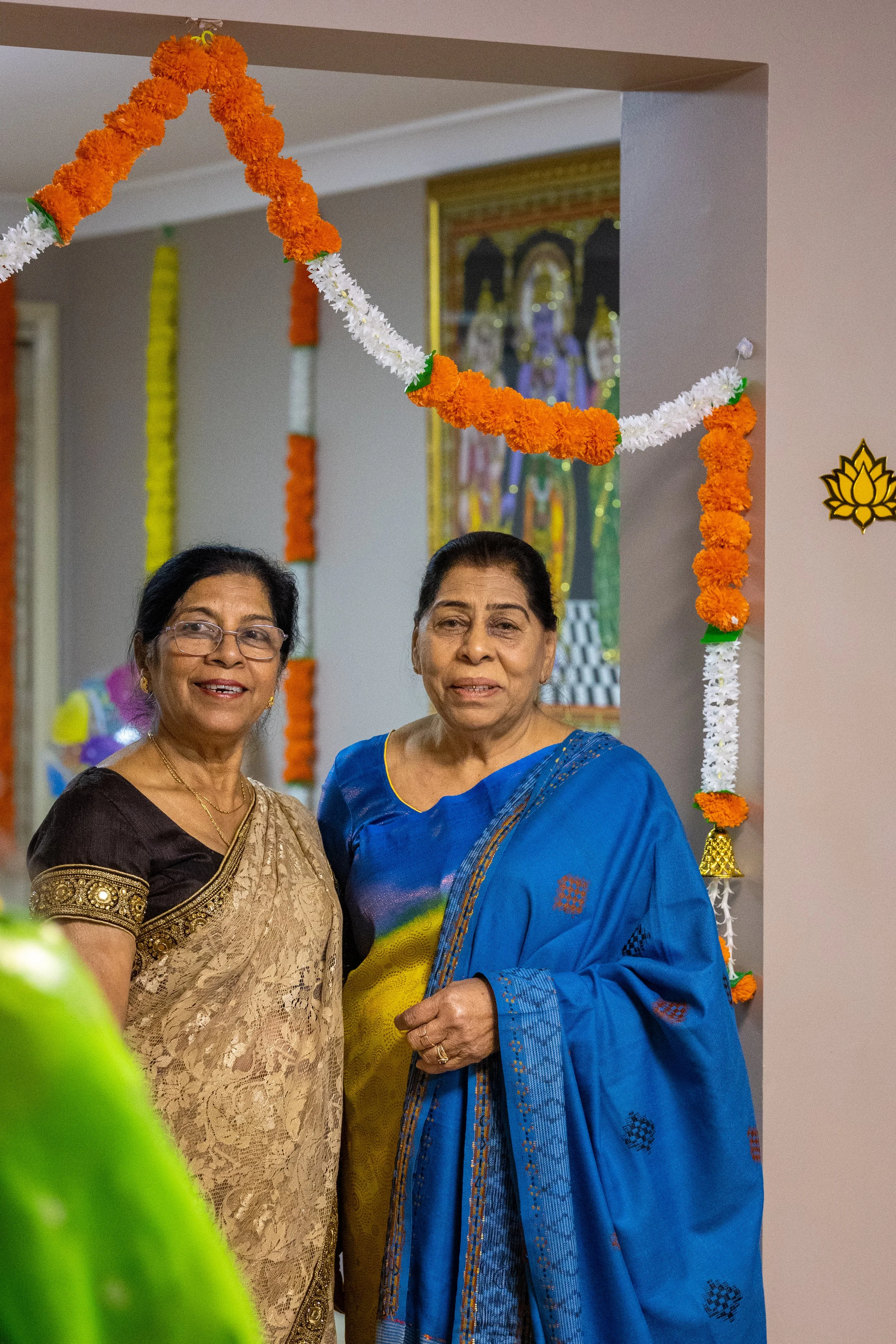 Two women dressed in traditional Indian sarees standing under a decorated floral arch for celebration.