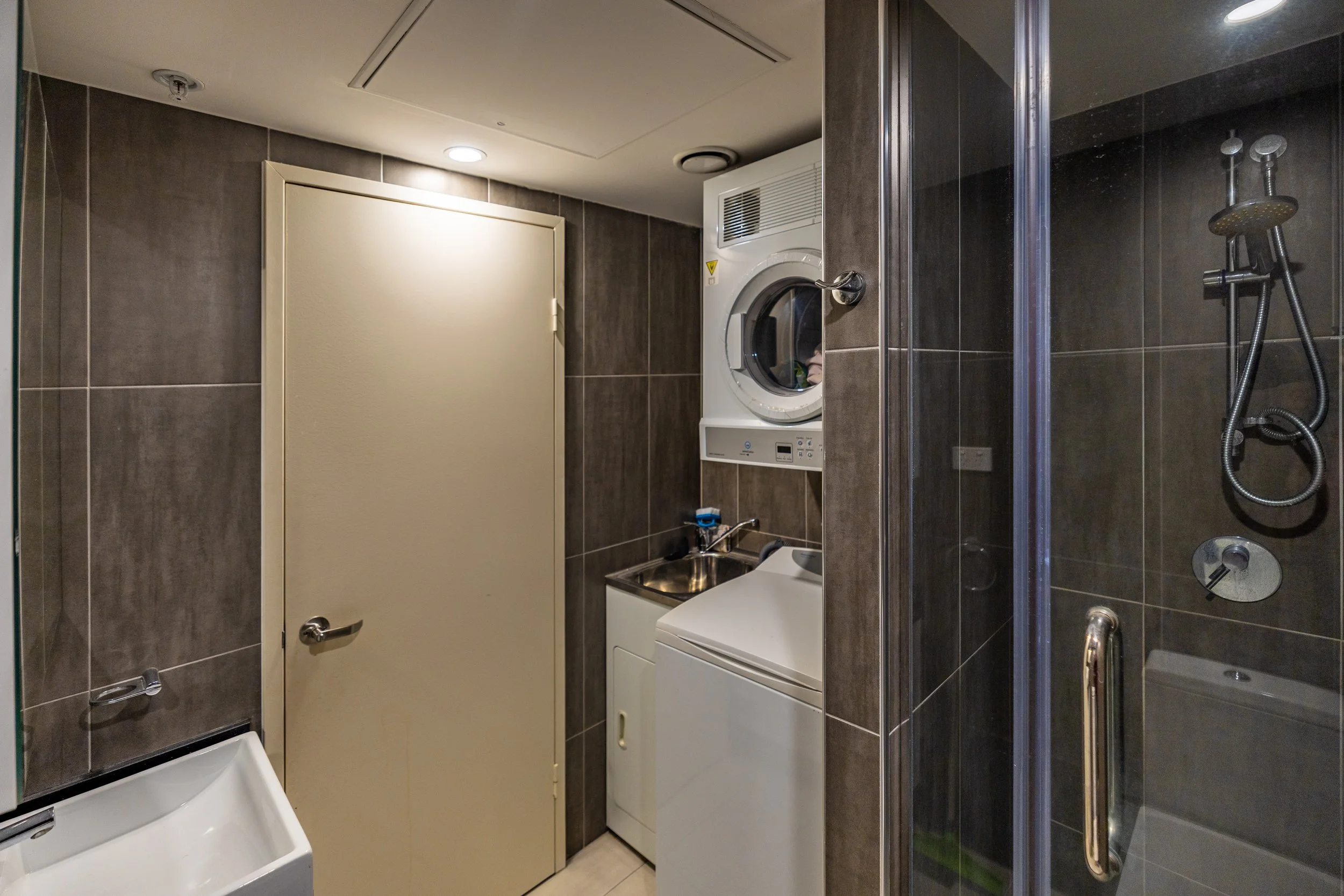 Small laundry room with beige door, dark brown tiled walls, washing machine, dryer, sink, and shower with glass door.