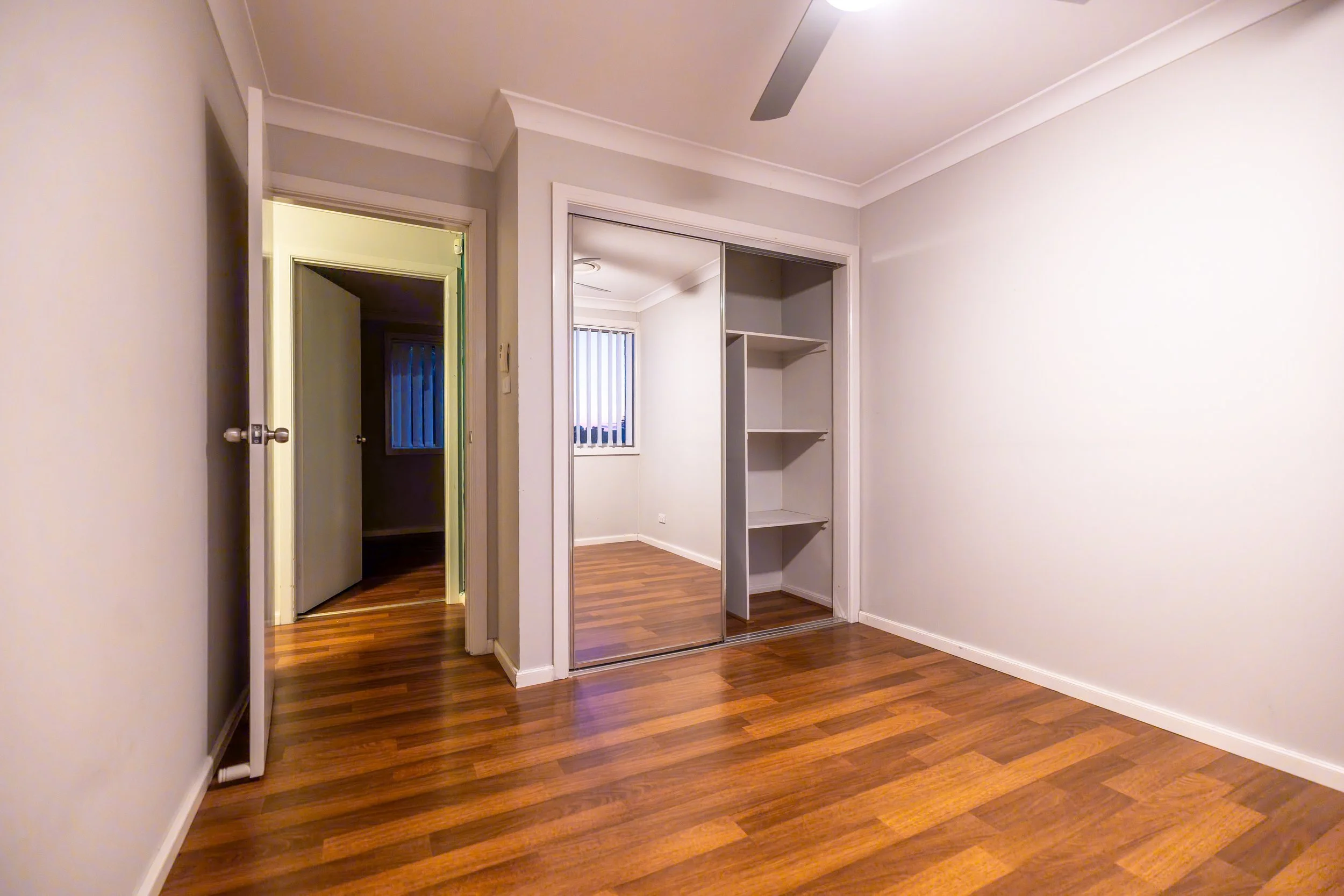 Empty bedroom with hardwood floors, white walls, a built-in closet with sliding mirror doors, and a doorway leading to a small room with a window with vertical blinds.