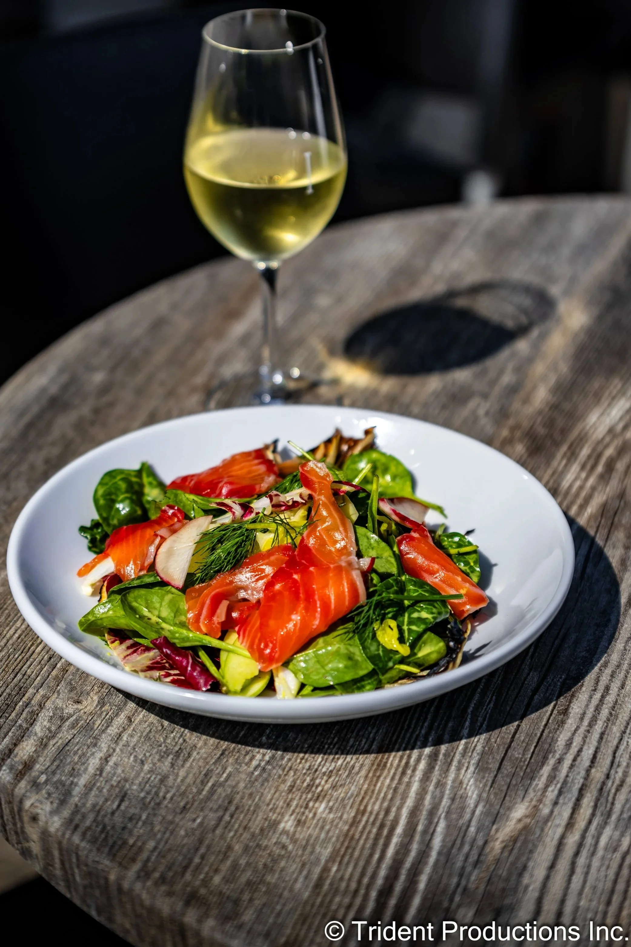 Salad with mixed greens and smoked salmon, served on a white plate, with a glass of white wine in the background on a wooden table