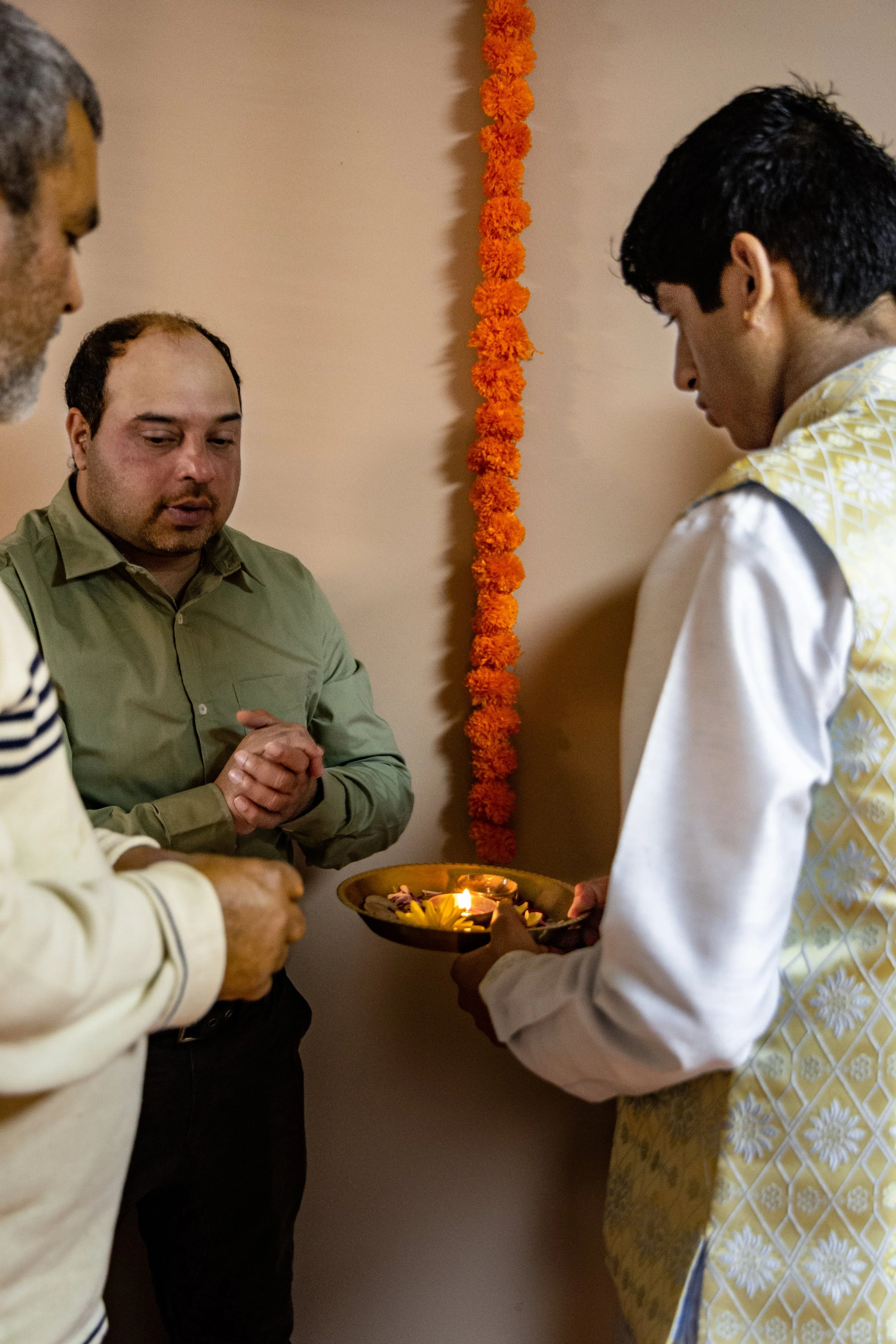 People engaged in a prayer ritual, one holding a traditional brass plate with lit candles and flower petals, during a religious ceremony, with marigold flower decorations on the wall.