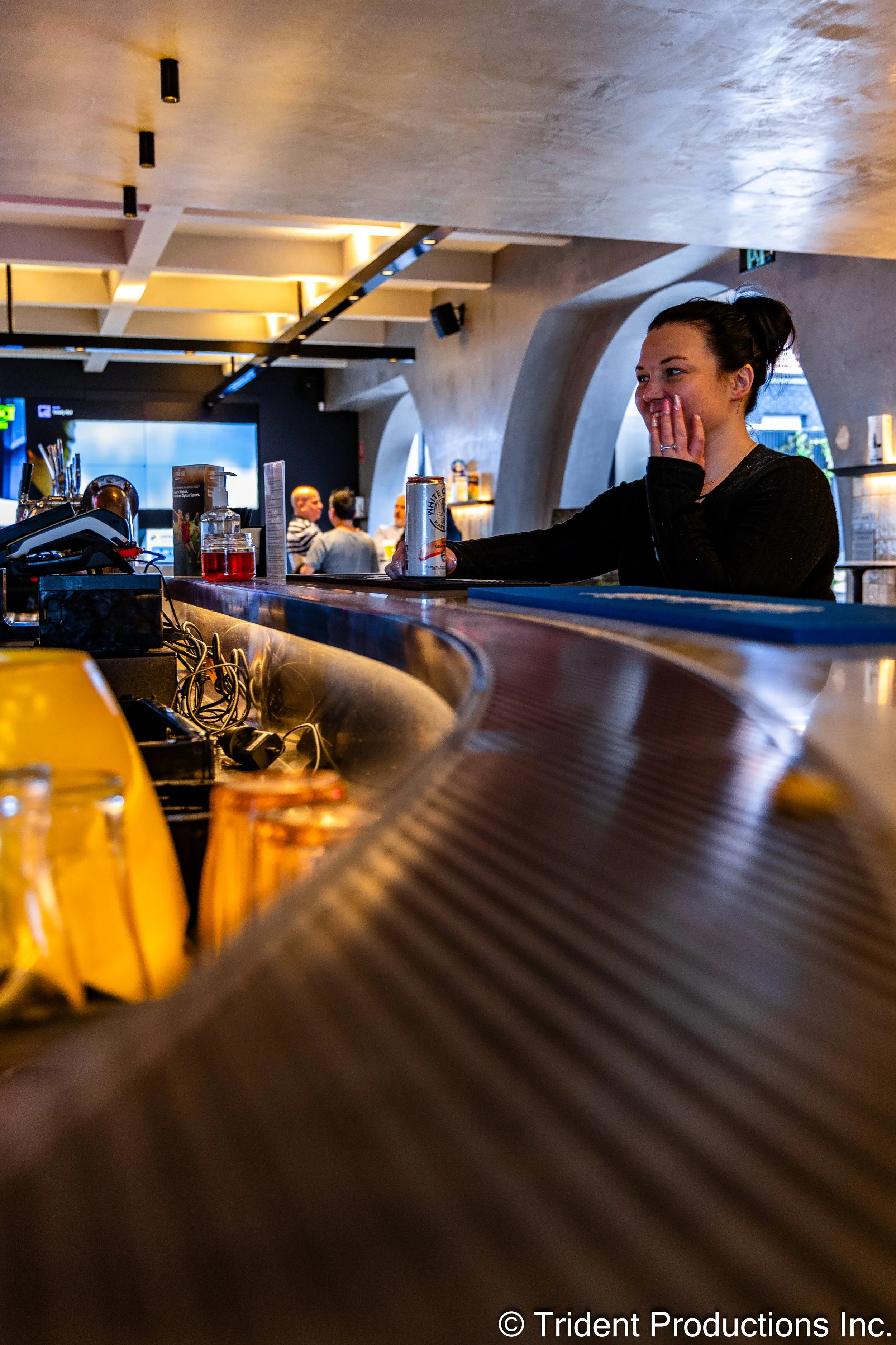 A woman with dark hair tied back, sitting at a bar counter in a modern, stylish bar or restaurant. She is smiling and holding a can of beverage, with her hand covering her mouth. The background shows a few other patrons, a large screen, and contempor