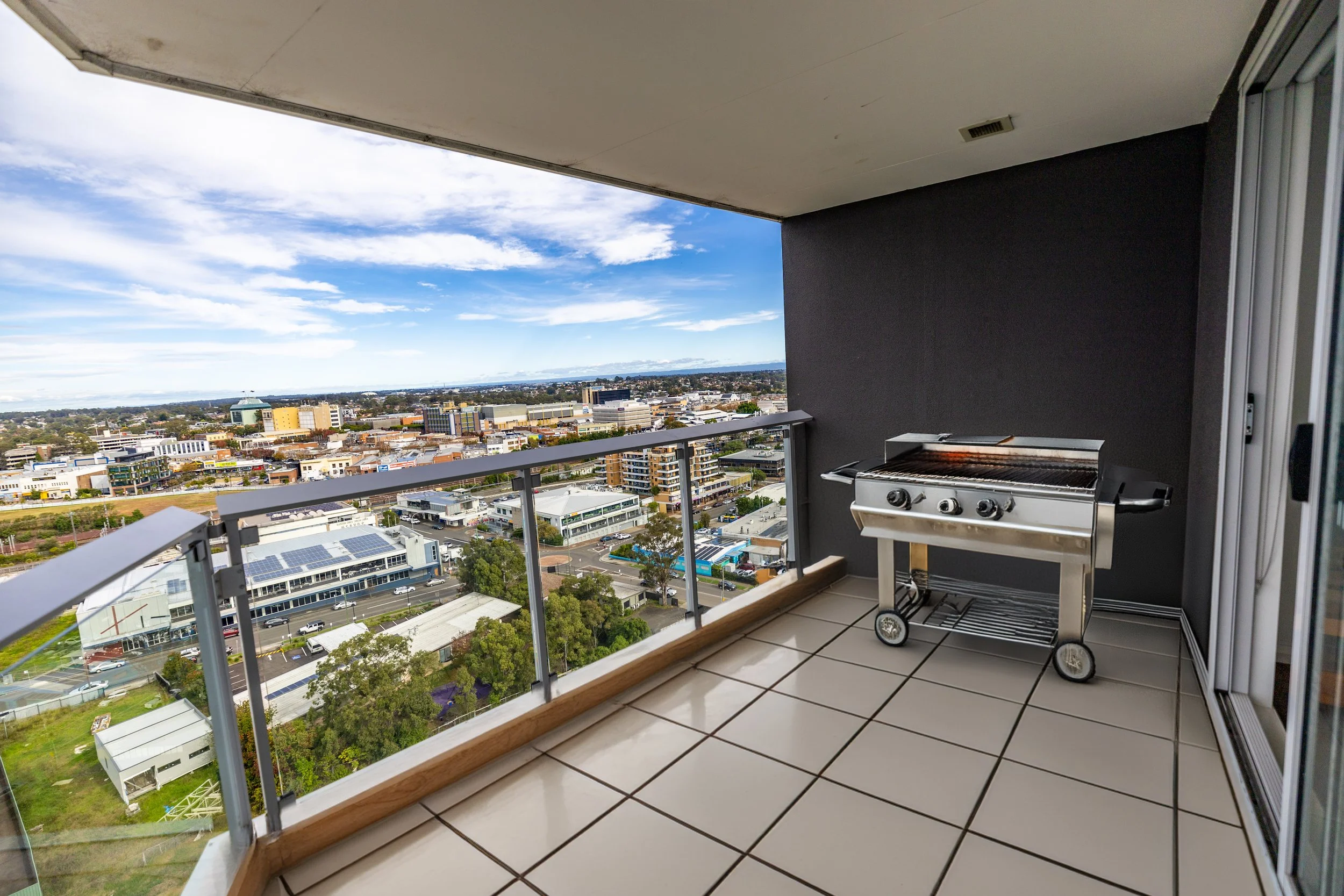 Balcony with a barbecue grill overlooking a cityscape with buildings, roads, and trees under a partly cloudy sky.