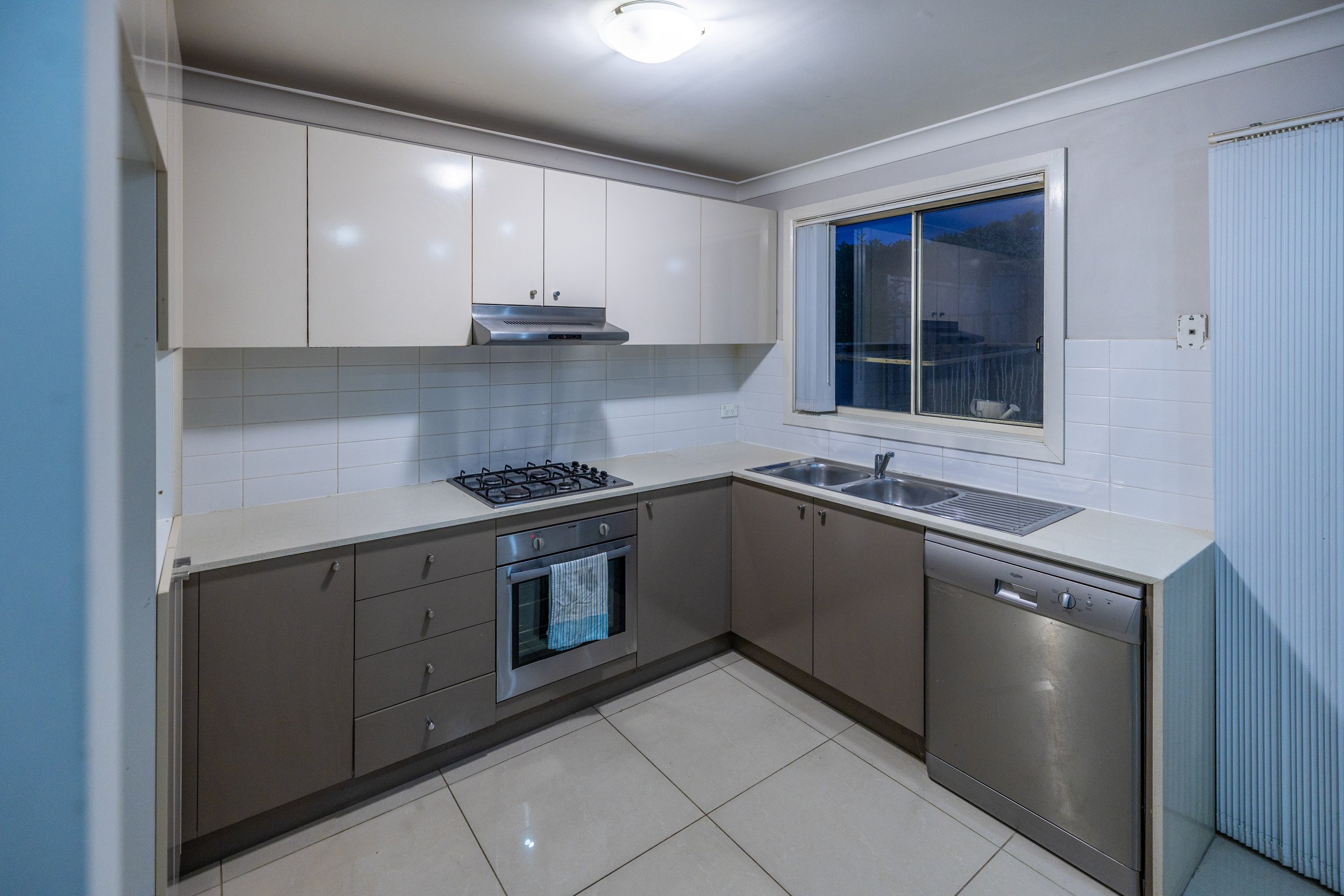 Kitchen with white upper cabinets, brown lower cabinets, stainless steel oven, gas stove, stainless steel dishwasher, double sink, and window above the sink.