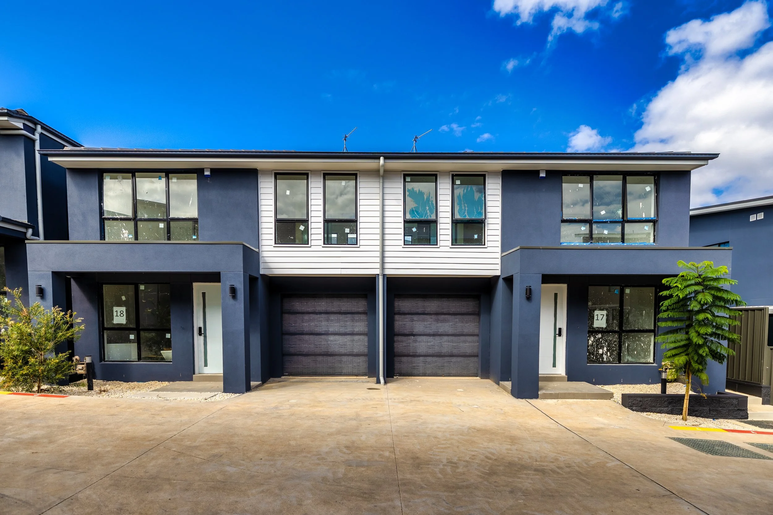 Modern multi-story duplex with dark blue and white exterior, large windows, and attached garages, under a partly cloudy sky.