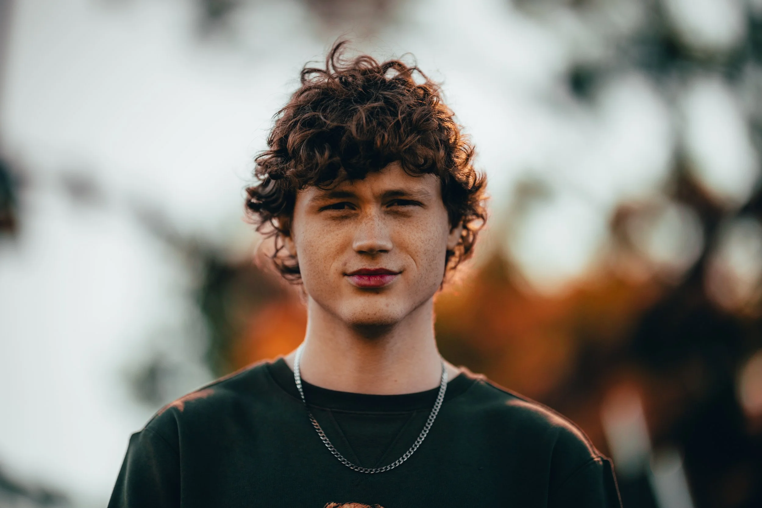 A young man with curly brown hair, light skin with freckles, wearing a black shirt and silver chains, standing outdoors with a blurred background of trees and orange leaves.