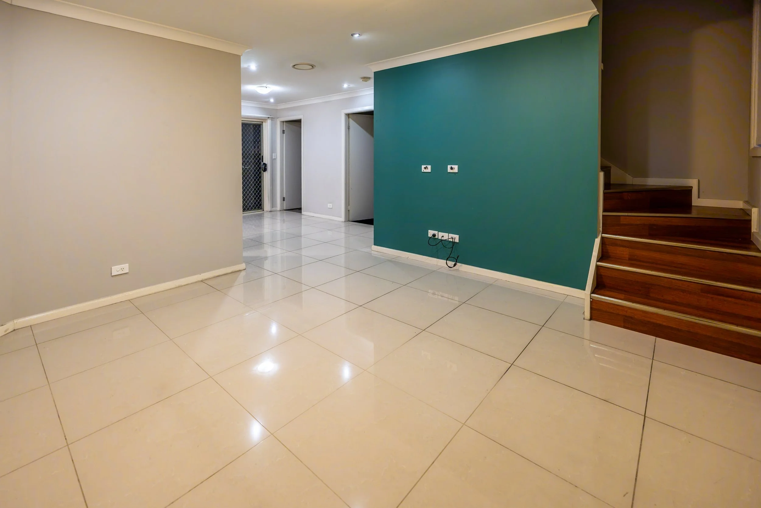 Empty living room with tiled floor, beige and dark teal walls, staircase to the right, and an entryway with a door and screen door in the background.