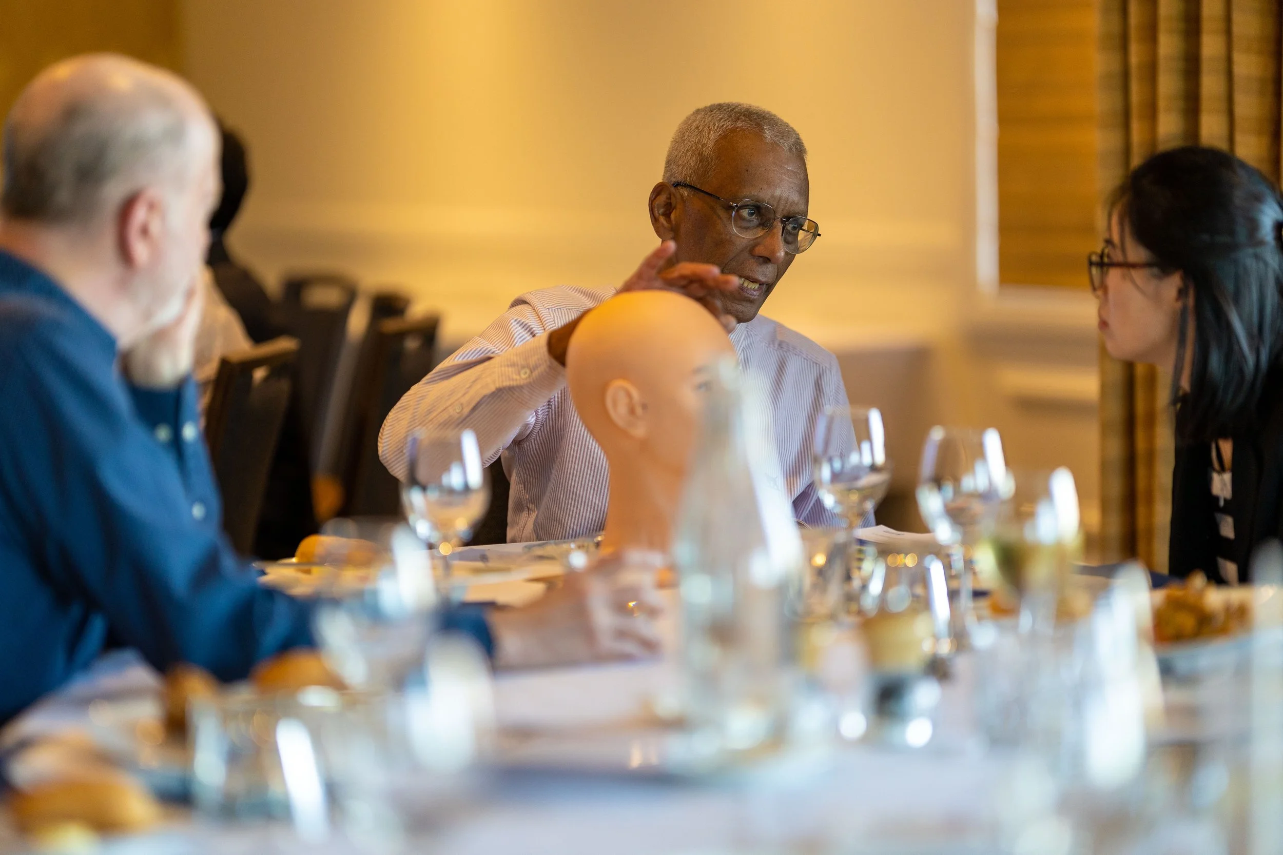 Three people seated at a dining table engaged in conversation, with glasses and plates on the table, in a well-lit room with yellow walls and window curtains.