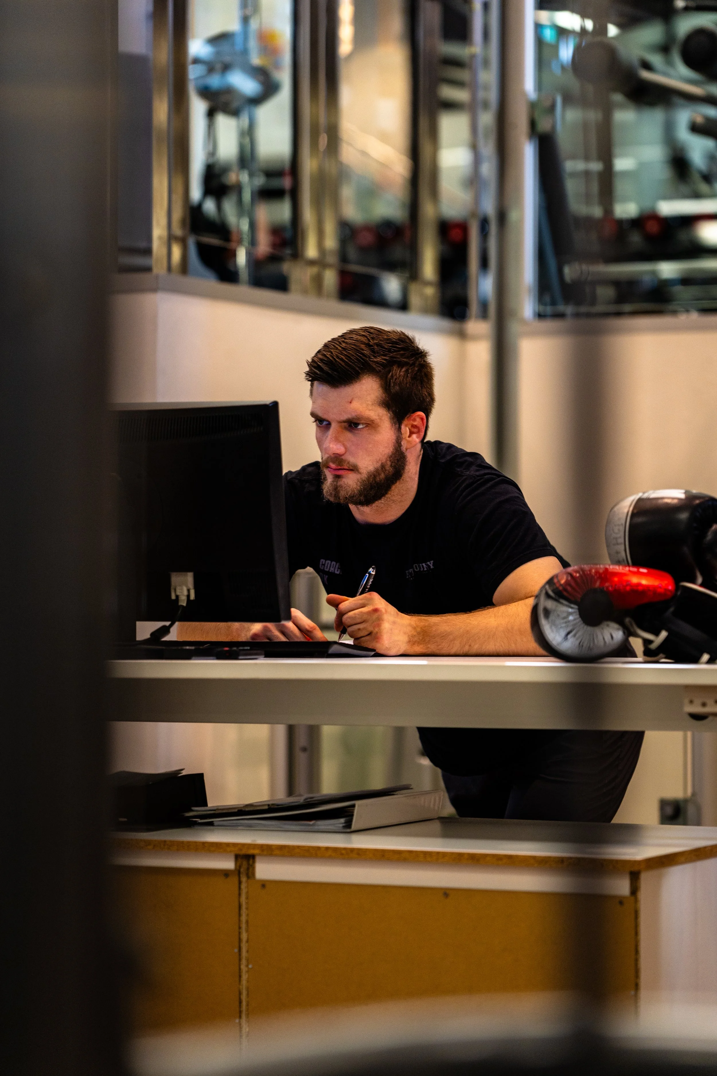 A man with a beard and short hair sitting at a desk, working on a computer, with boxing gloves on the desk in a gym or training facility.