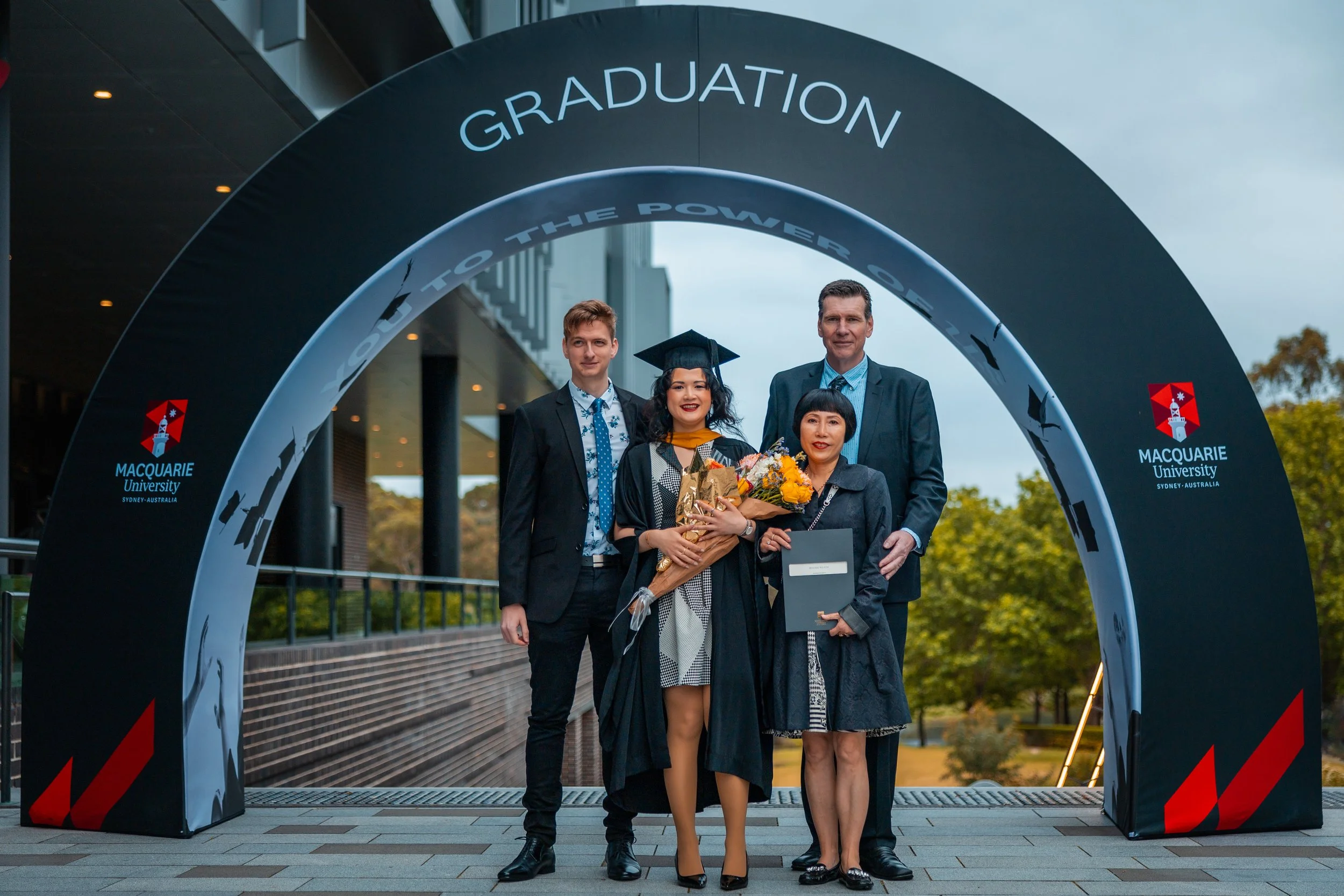A graduation ceremony at Macquarie University in Sydney, Australia, with four people standing under a large arch that reads 'Graduation'. One woman is wearing a cap and gown, holding a bouquet of flowers and a diploma, flanked by a man and two women 
