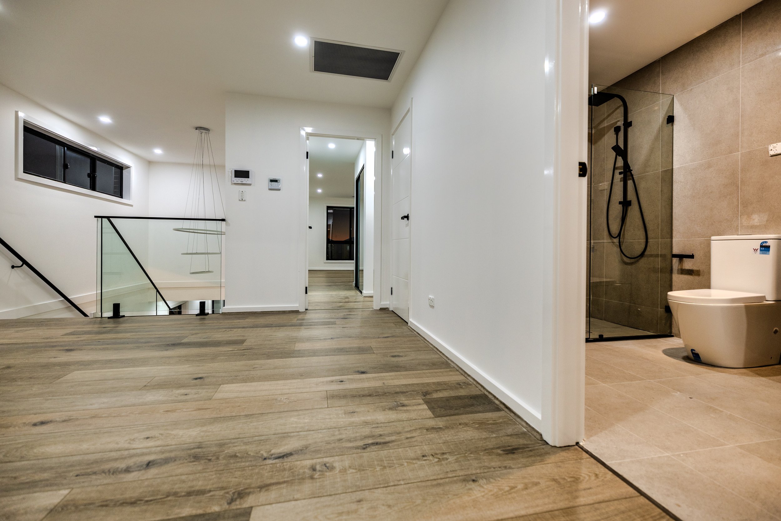 Modern interior home hallway with wooden flooring leading to a bathroom with glass shower and toilet, white walls, multiple ceiling lights, and a staircase with glass railing.