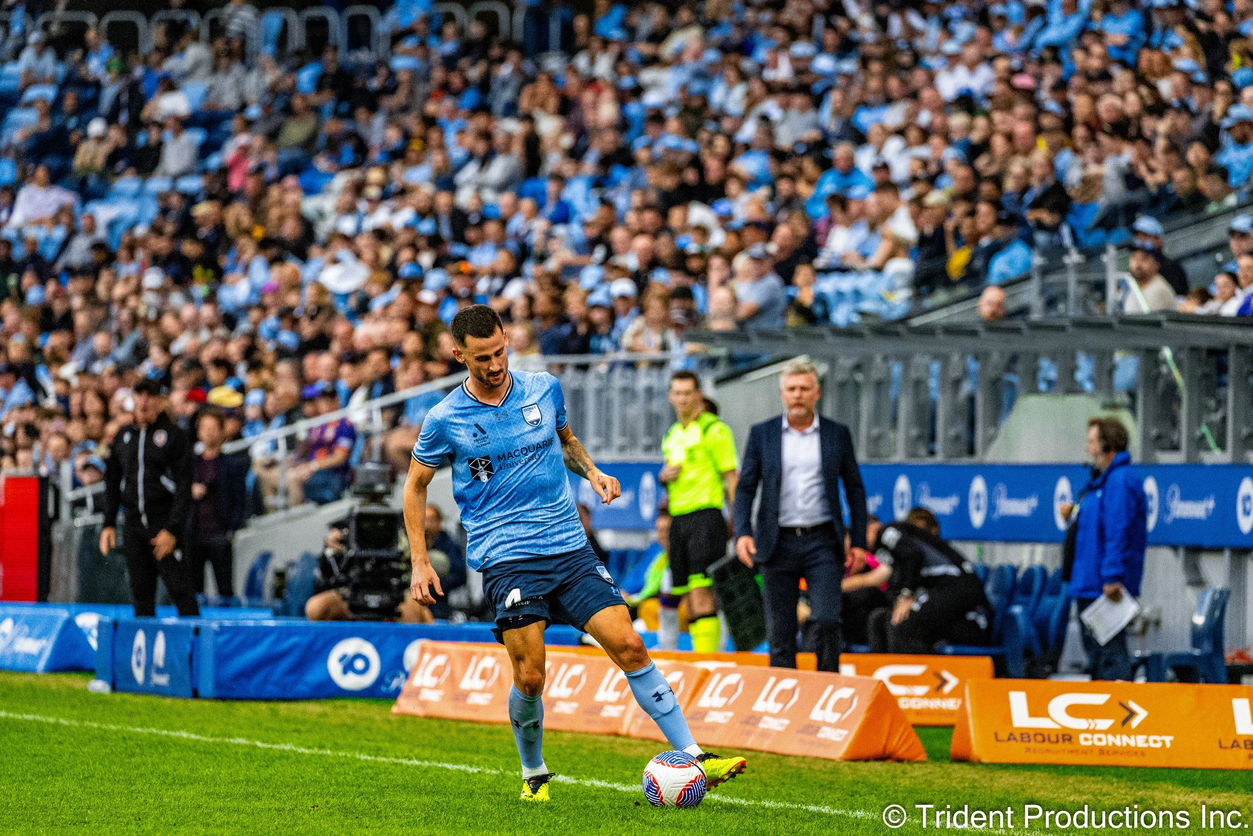 Soccer player in light blue uniform kicking a soccer ball on field with crowd in stands and officials watching in background.