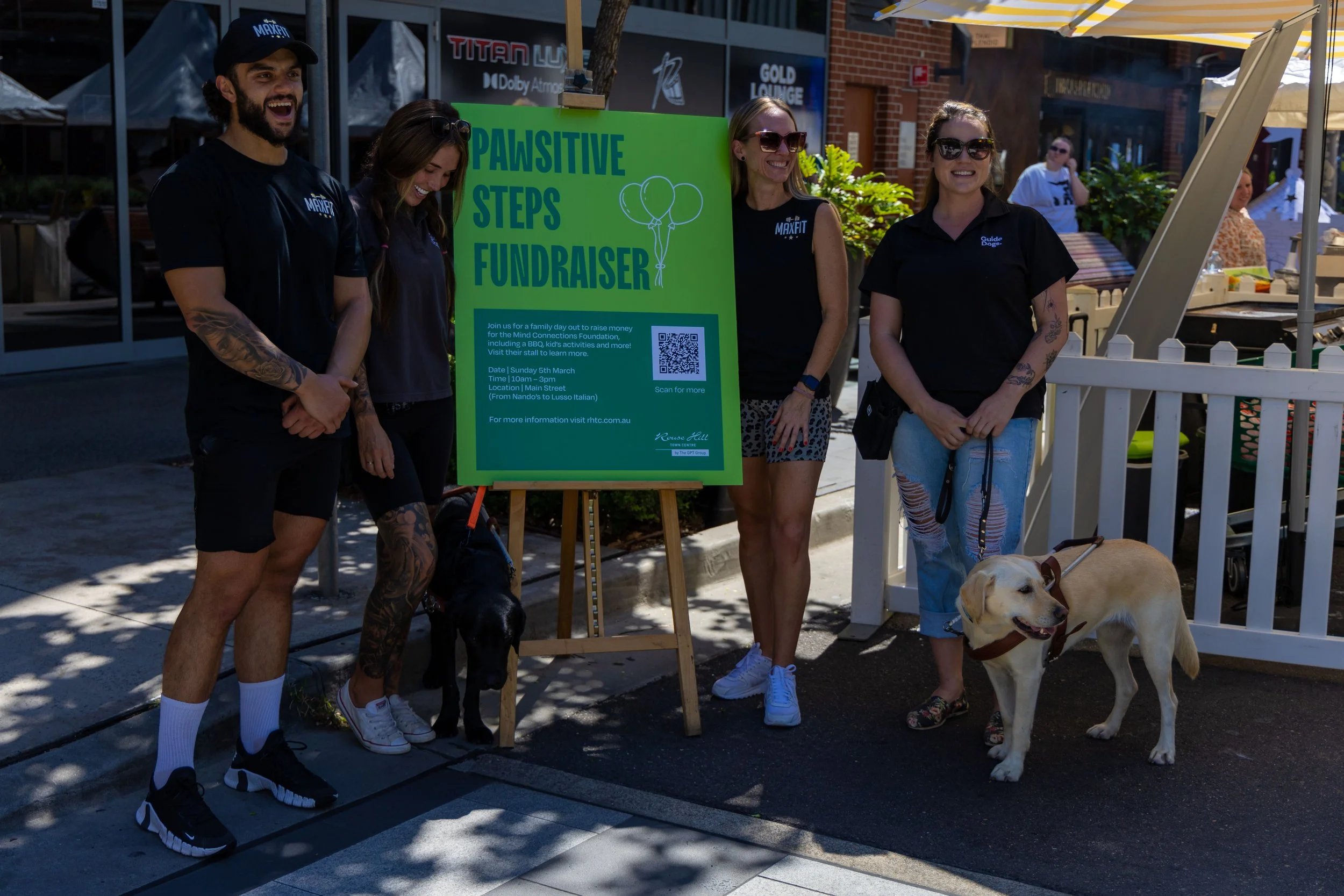 Group of people standing outdoors next to a green sign that reads 'Pawsitive Steps Fundraiser' with balloons, at a family event for charity, with dogs and food stalls in the background.