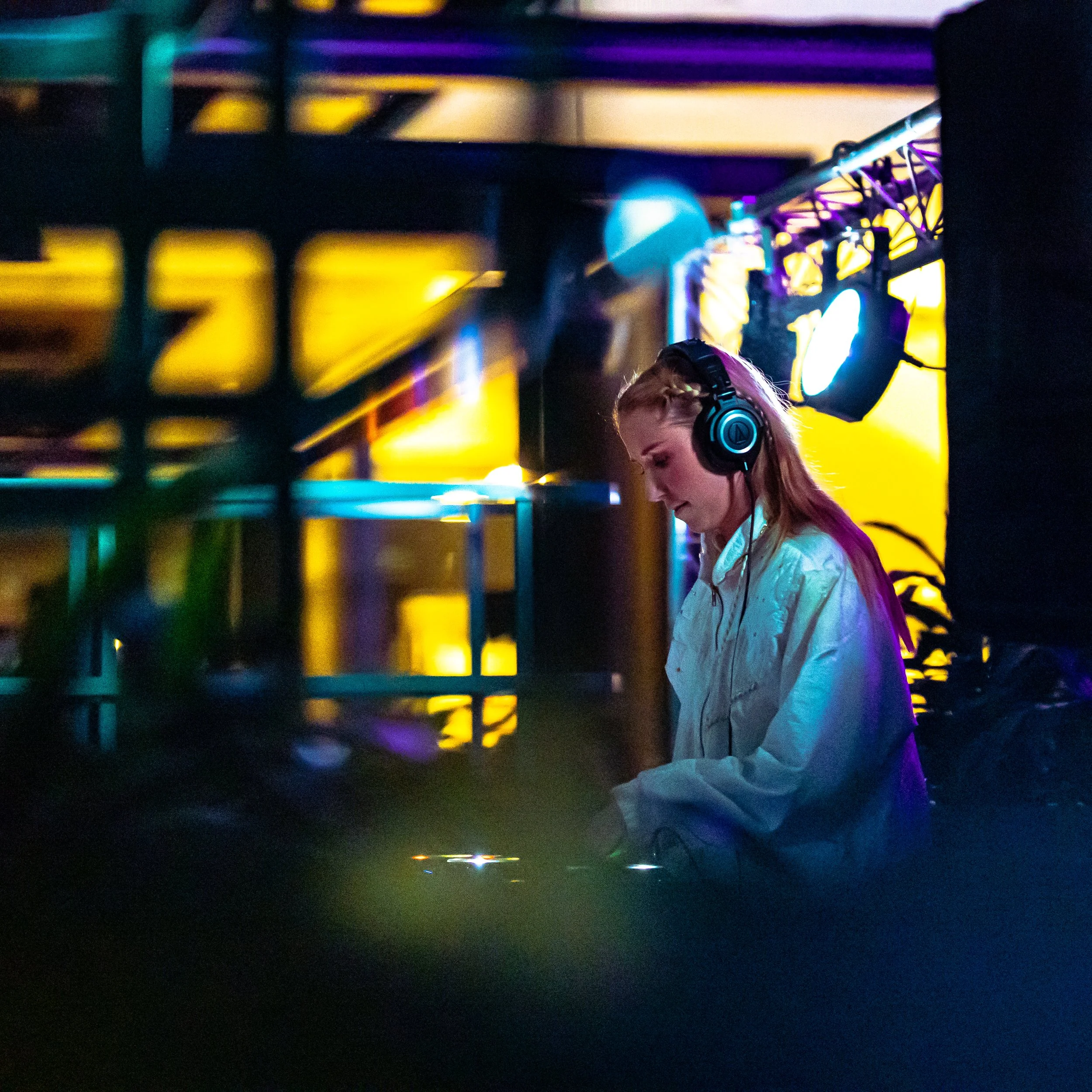 A woman with long blonde hair wearing headphones and a white shirt, DJing in a colorful, dimly lit nightclub or event space.