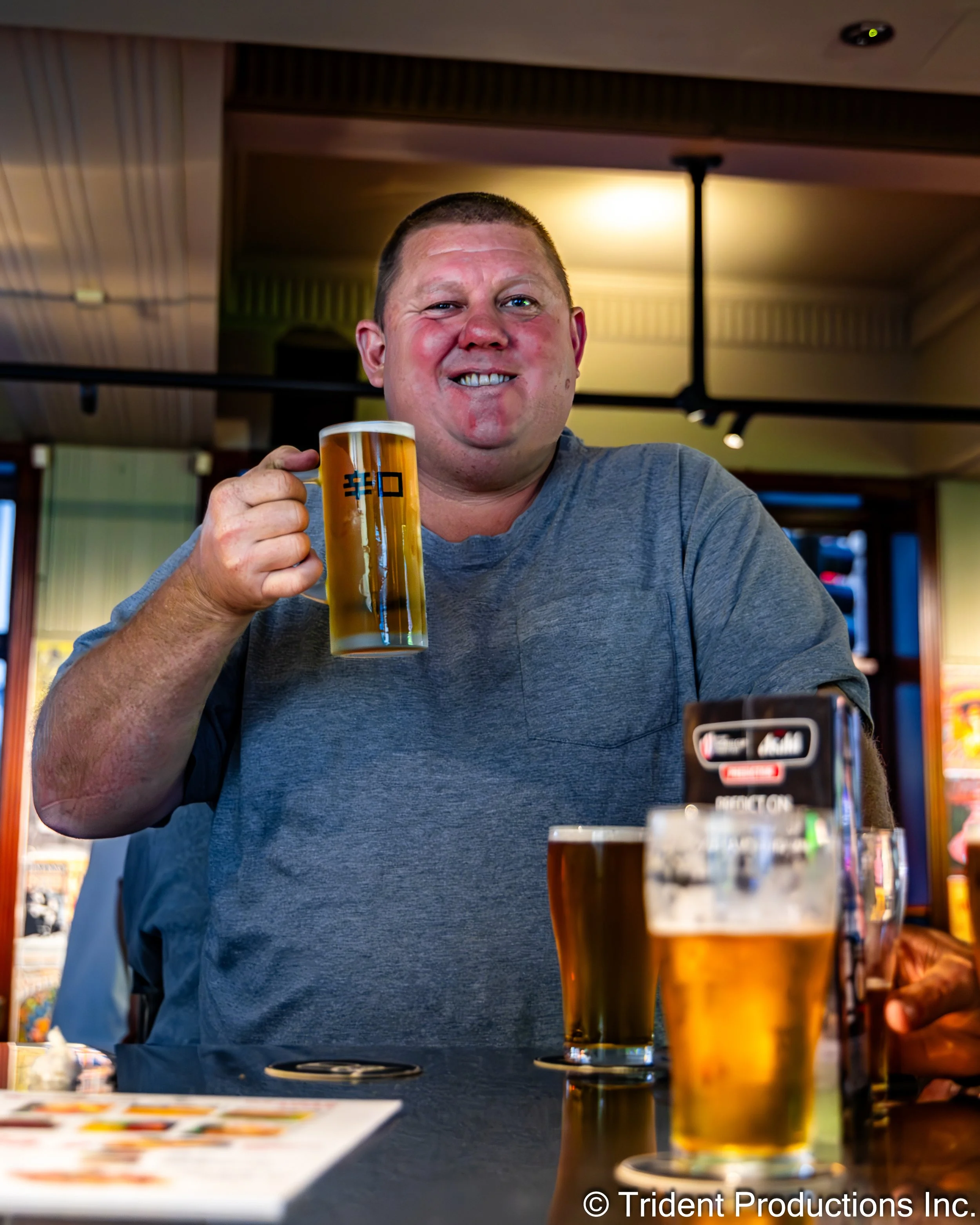 A man in a gray t-shirt holding a glass of beer in a bar or pub setting, smiling at the camera, with other glasses of beer on the table in front of him.