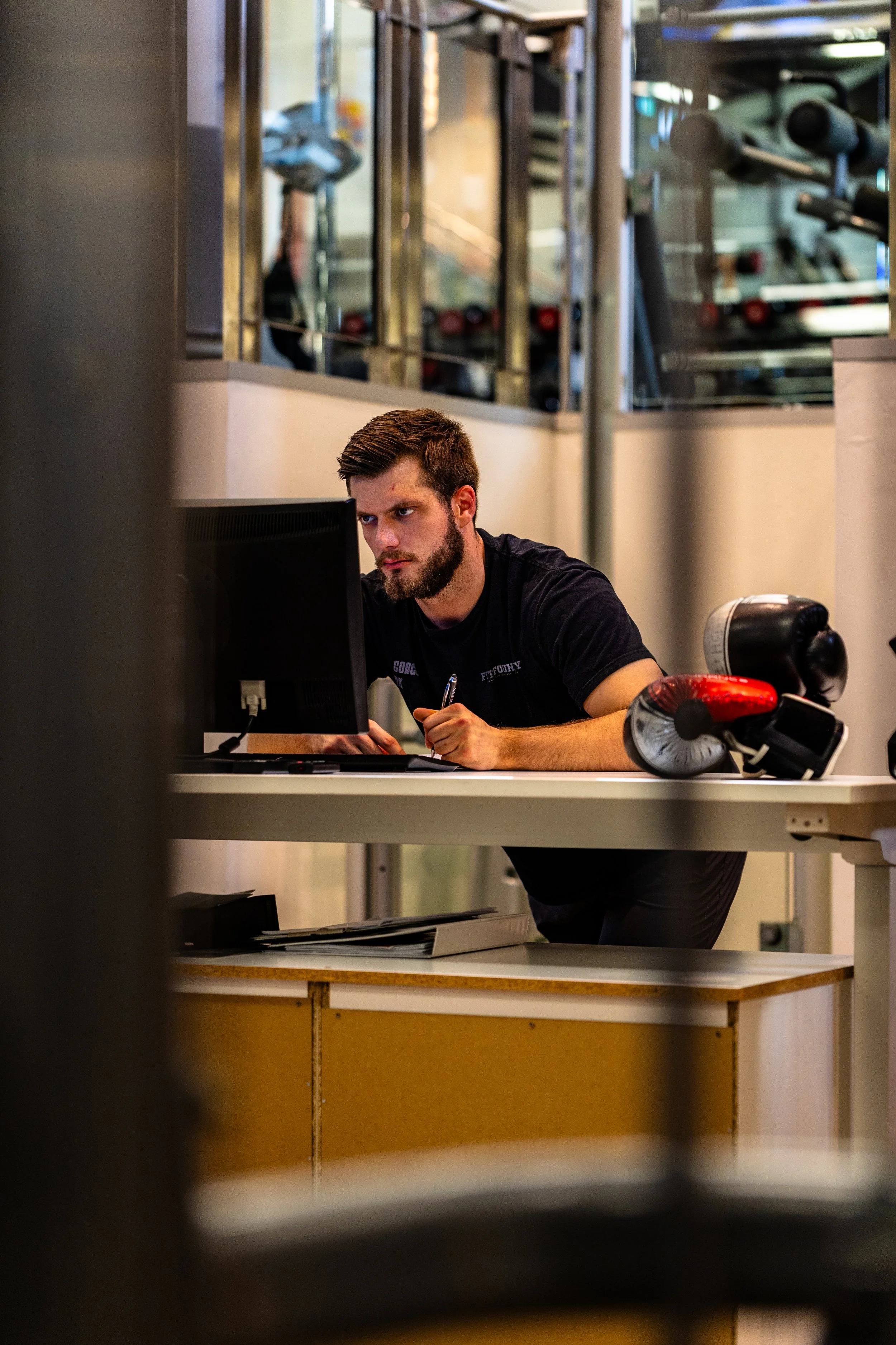 A man working on a computer at a desk in a gym, with boxing gloves and a punching bag nearby.