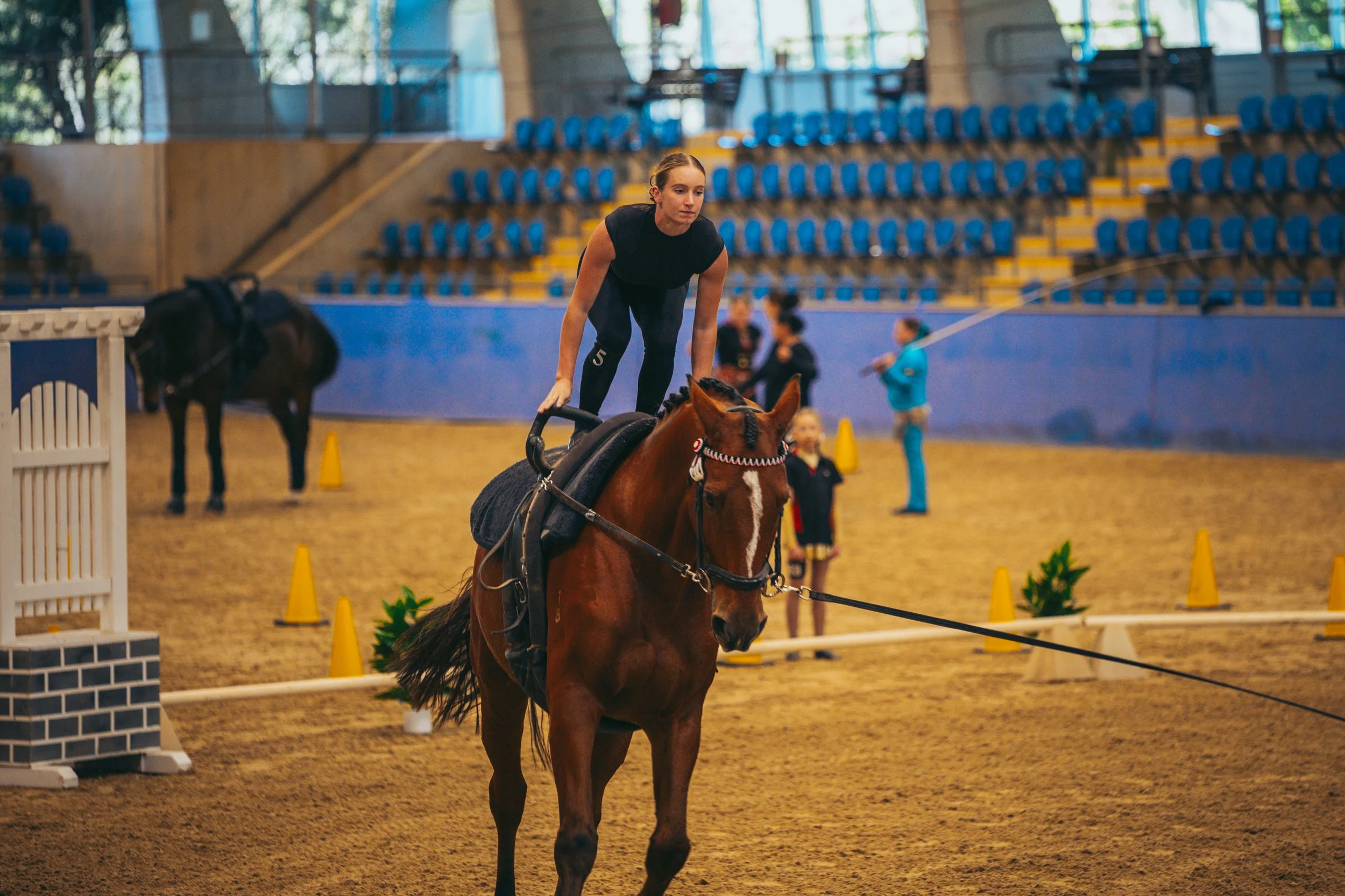 A woman riding a brown horse in an indoor equestrian arena, practicing dressage, with spectators sitting in blue seats and other people and horses in the background.