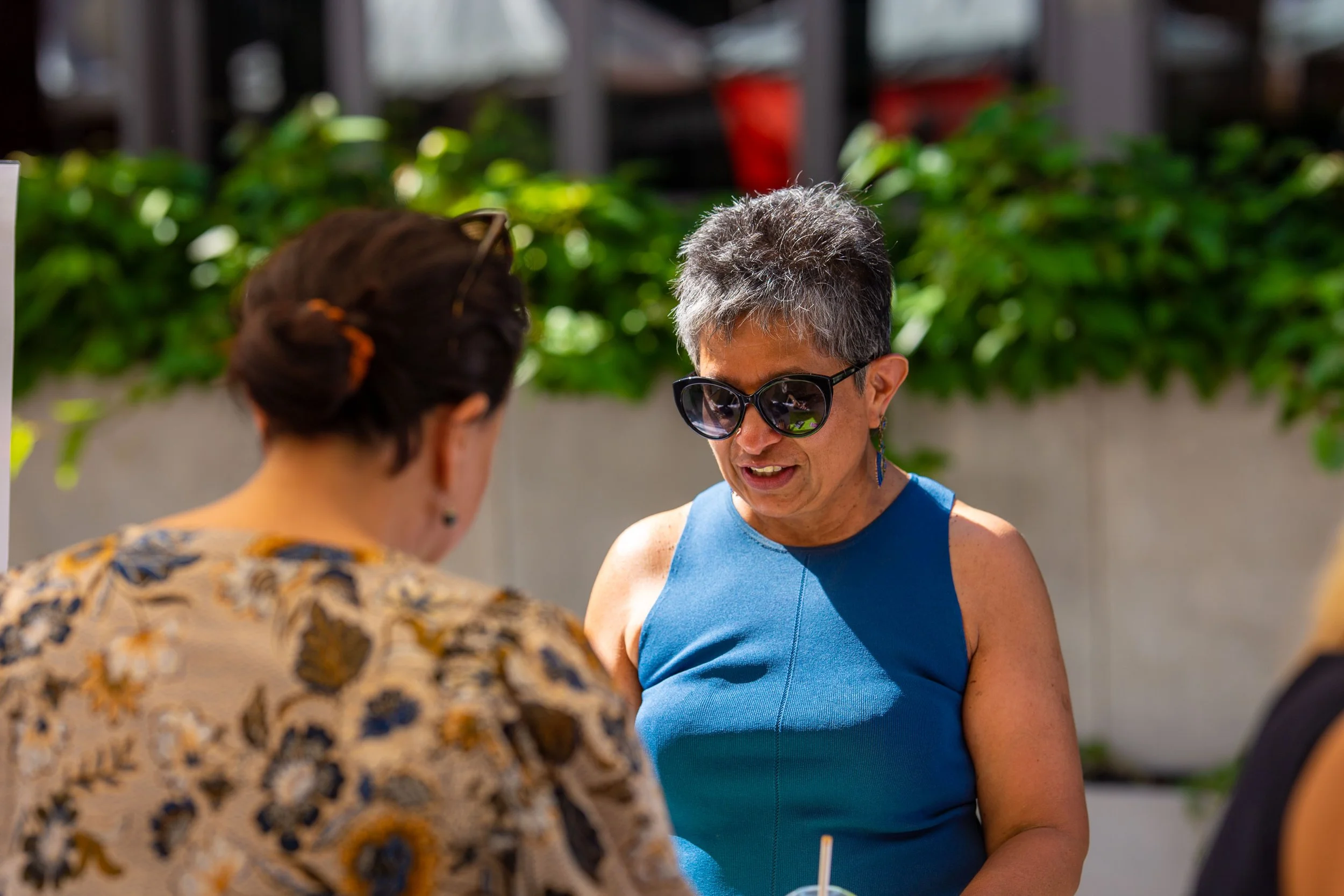 A woman with short gray hair wearing large sunglasses and a sleeveless blue top, smiling and talking to another woman with dark hair and glasses, outdoors with greenery in the background.