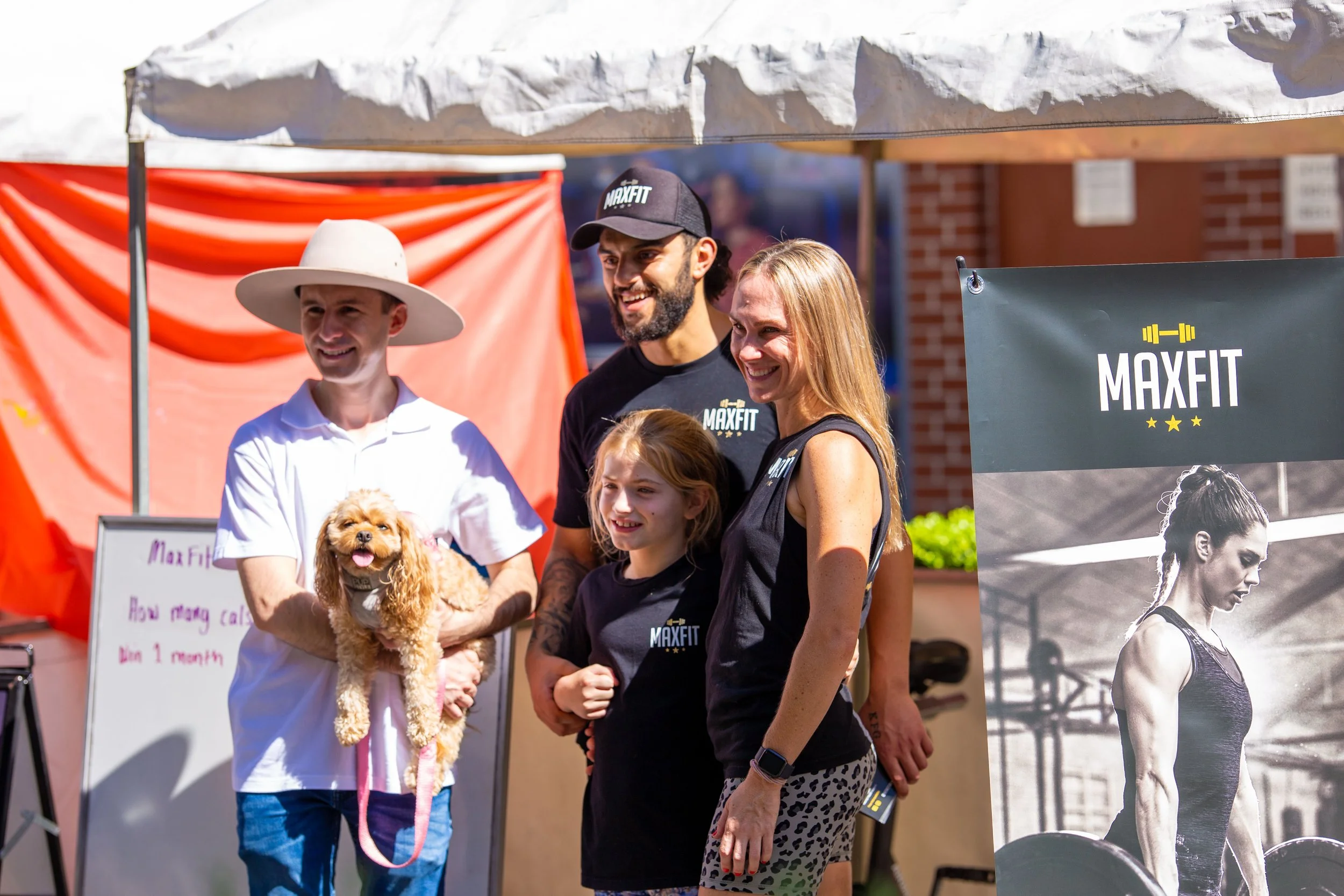 A group photograph of five people and a dog at an outdoor fitness event, with a MaxFit banner in the background. The person on the far left is holding a small brown dog, wearing a white wide-brimmed hat and white polo shirt. Other individuals are wea