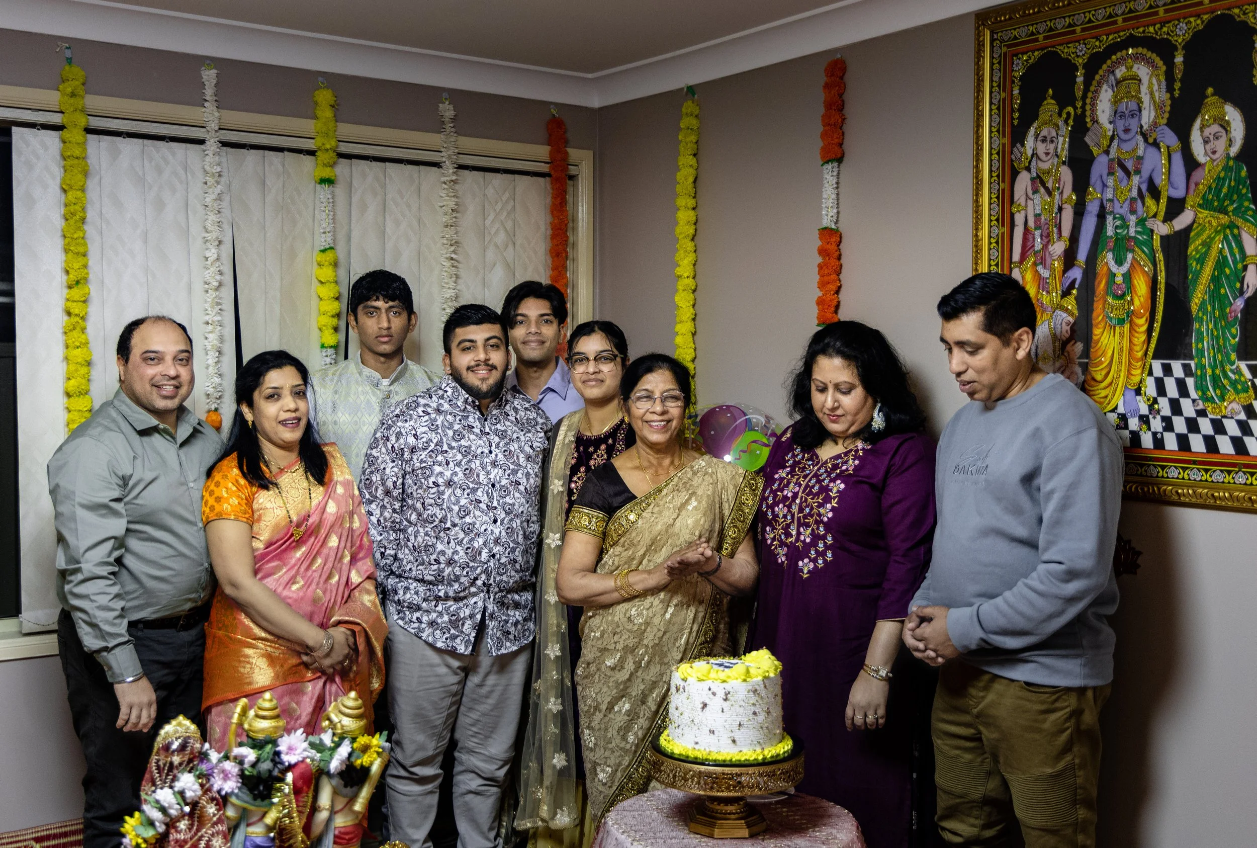 Family celebrating a traditional Indian birthday, with a cake, in front of a colorful portrait of Hindu deities, decorated with marigold and flower garlands.