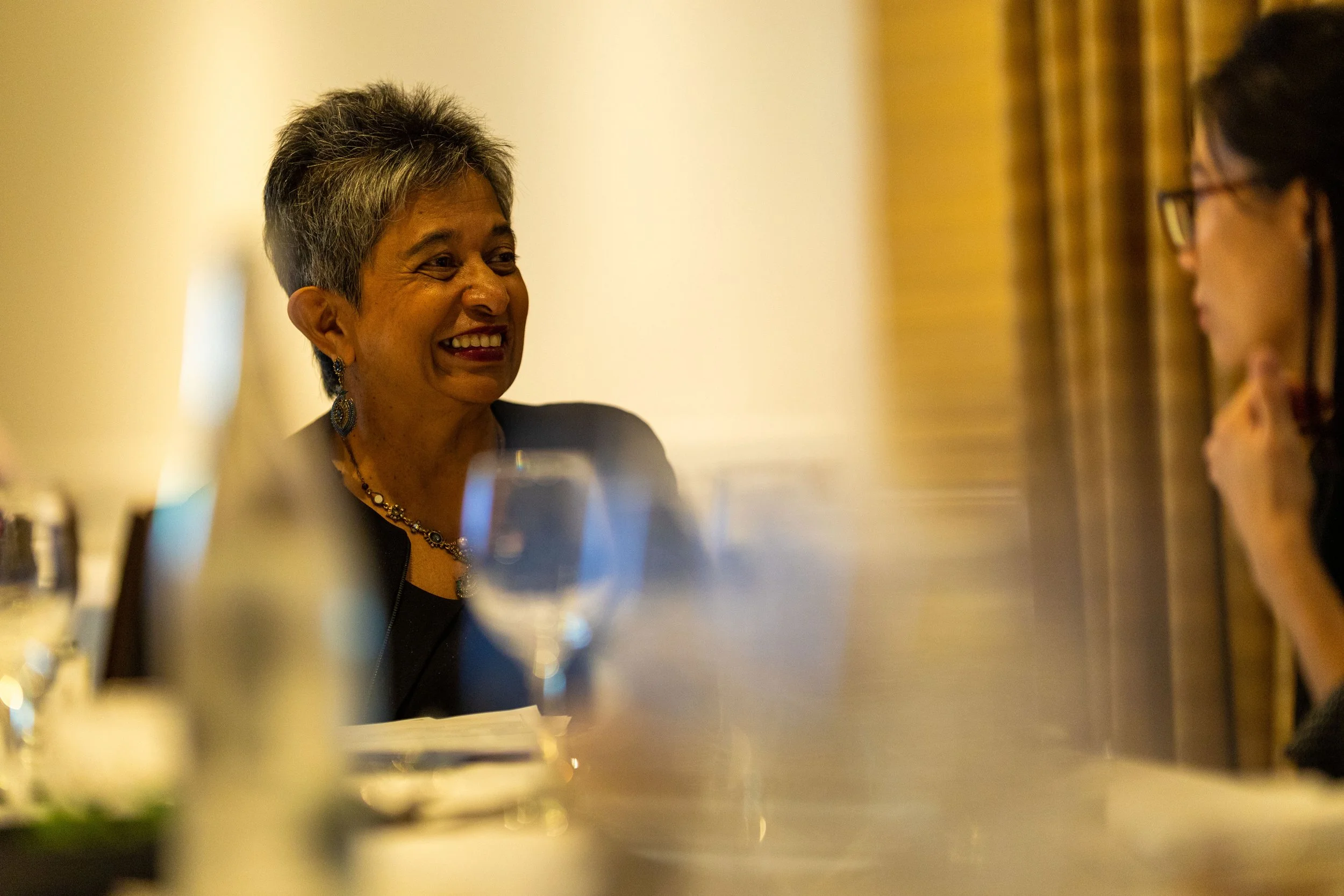 Two women sitting at a dinner table, talking and smiling, with glassware and table settings visible.