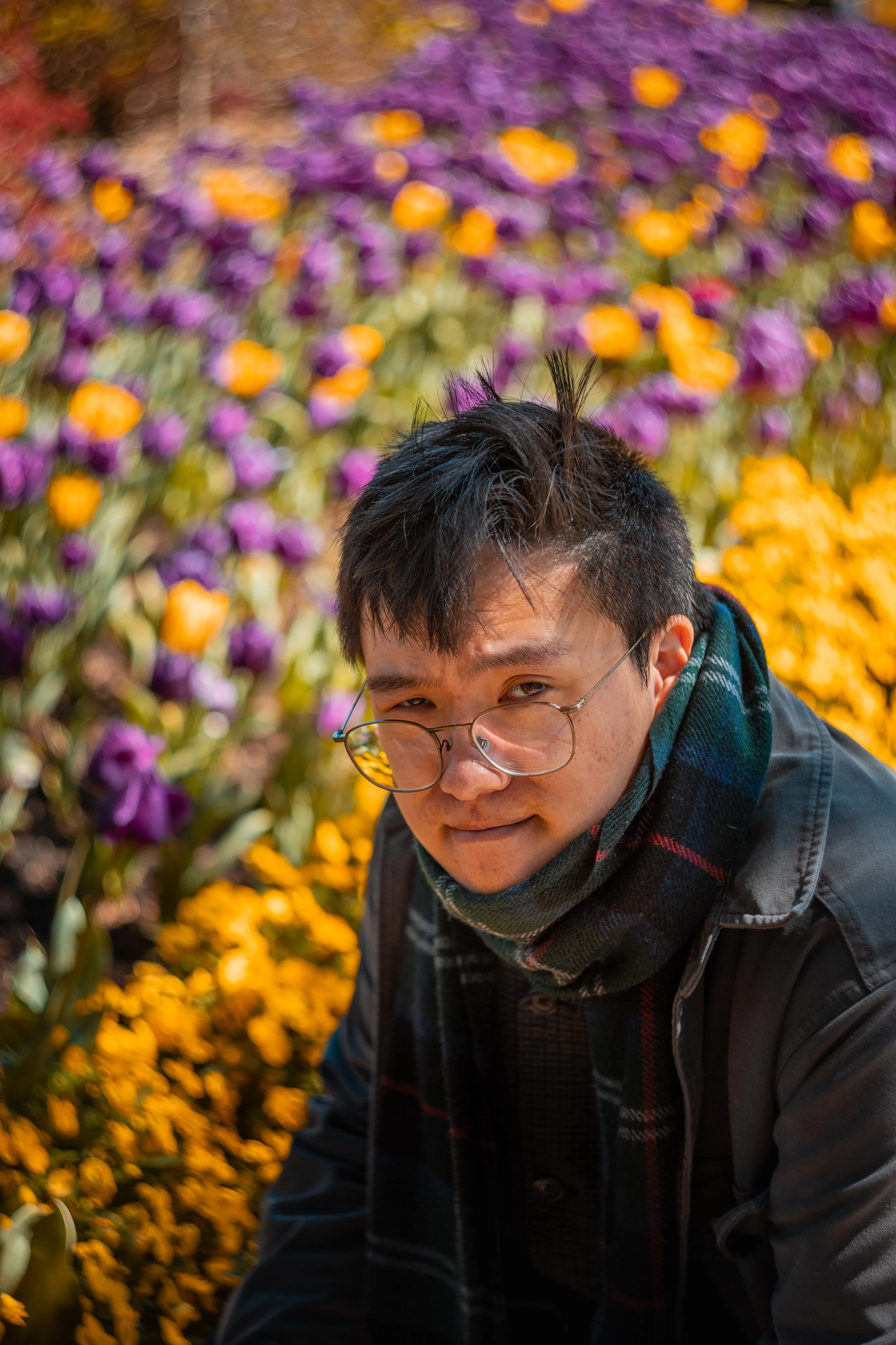 A young man with glasses, wearing a scarf and dark jacket, crouching among colorful tulips and flowers in a garden.