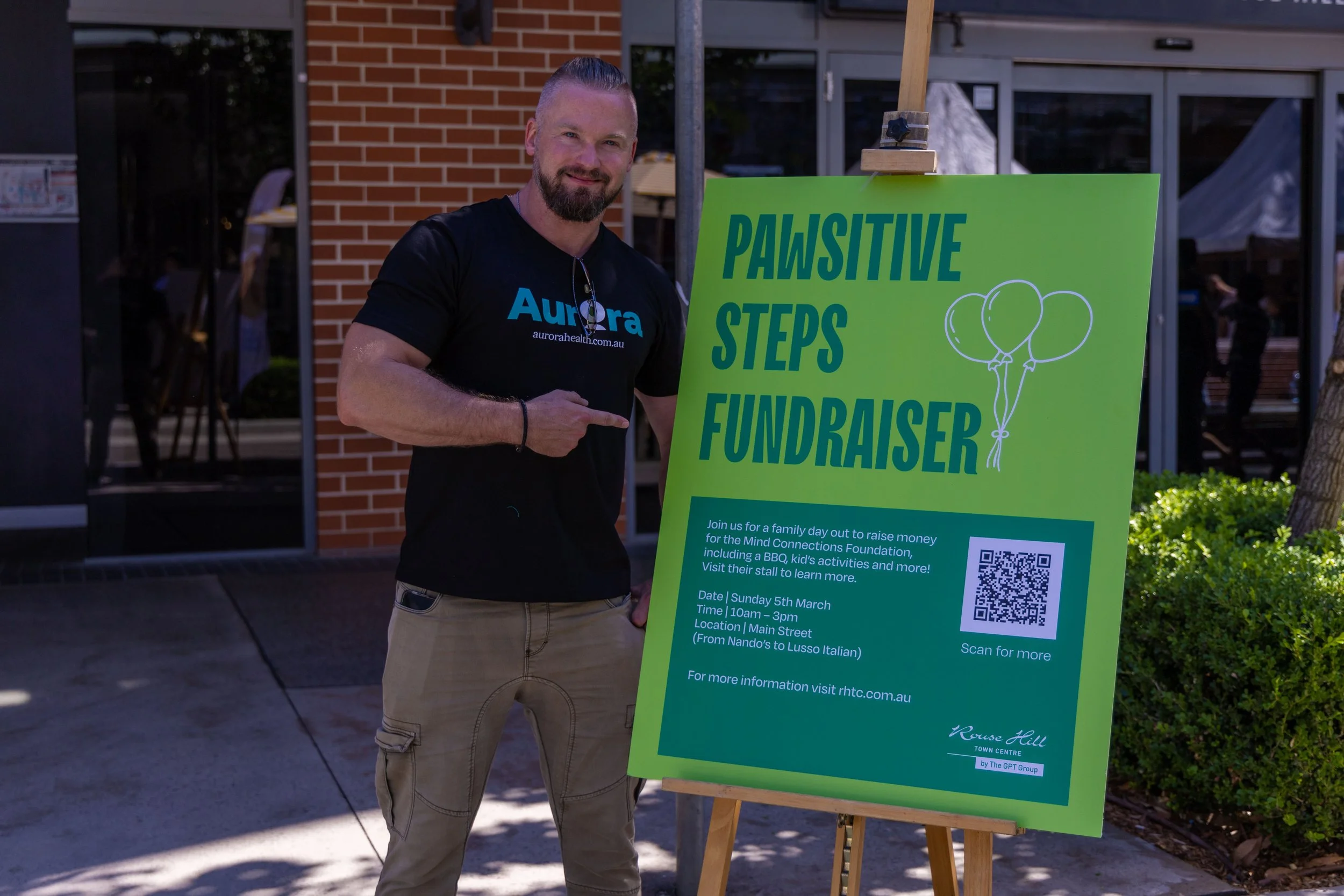 A man stands next to a bright green poster board with details about a fundraising event, pointing at the poster with his right hand. The poster mentions a family day event for the Mind Connections Foundation on Sunday, March 5th from 10 am to 3 pm on