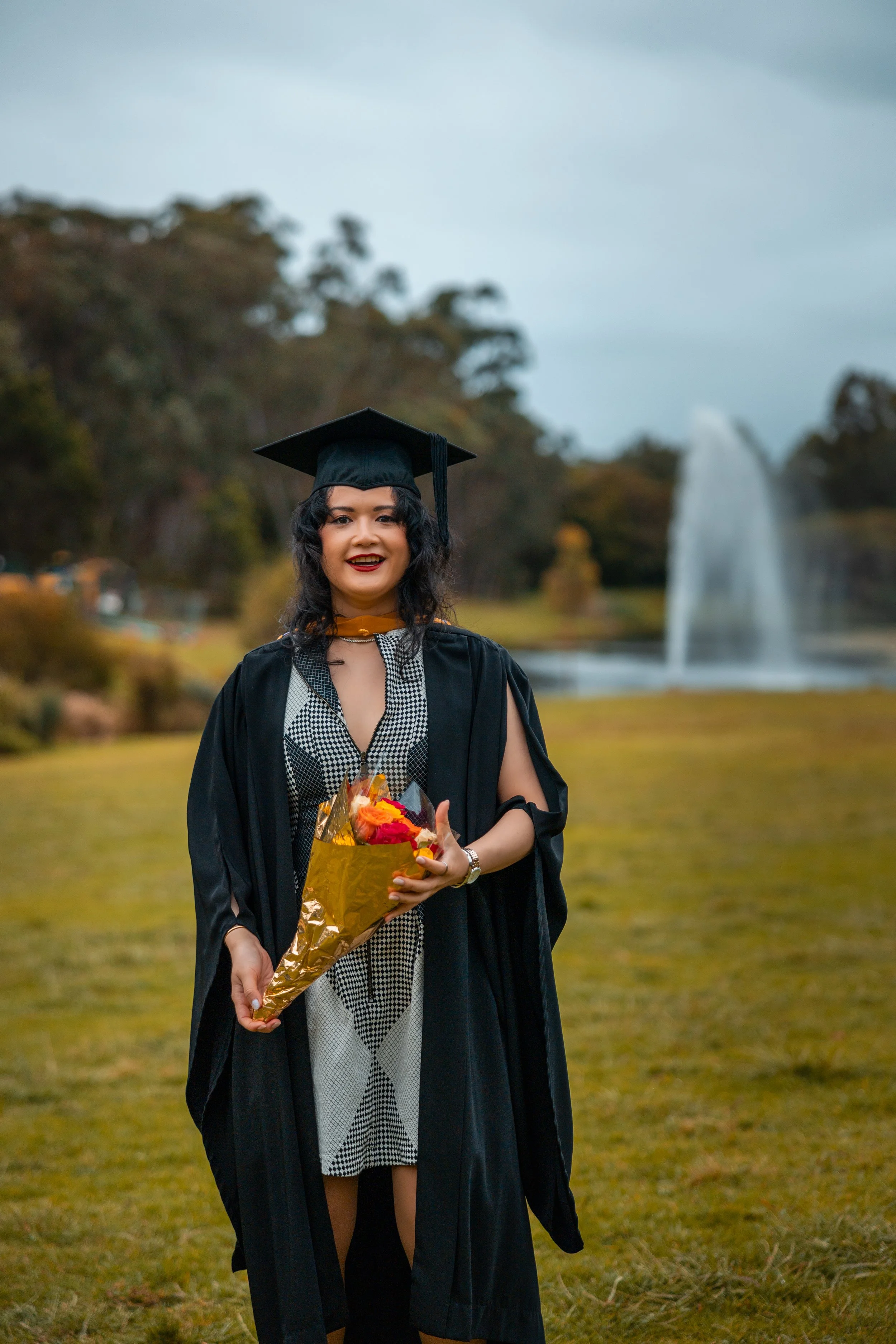 A woman in a graduation cap and gown holding a bouquet of flowers standing outdoors near a fountain and trees.