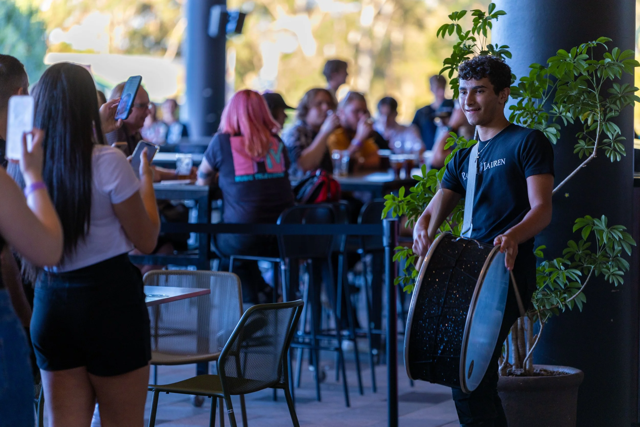 A young man holding a large circular musical instrument, possibly an African drum, near a potted plant, smiling, while people in the background are taking photos and socializing in a casual outdoor or semi-outdoor setting.