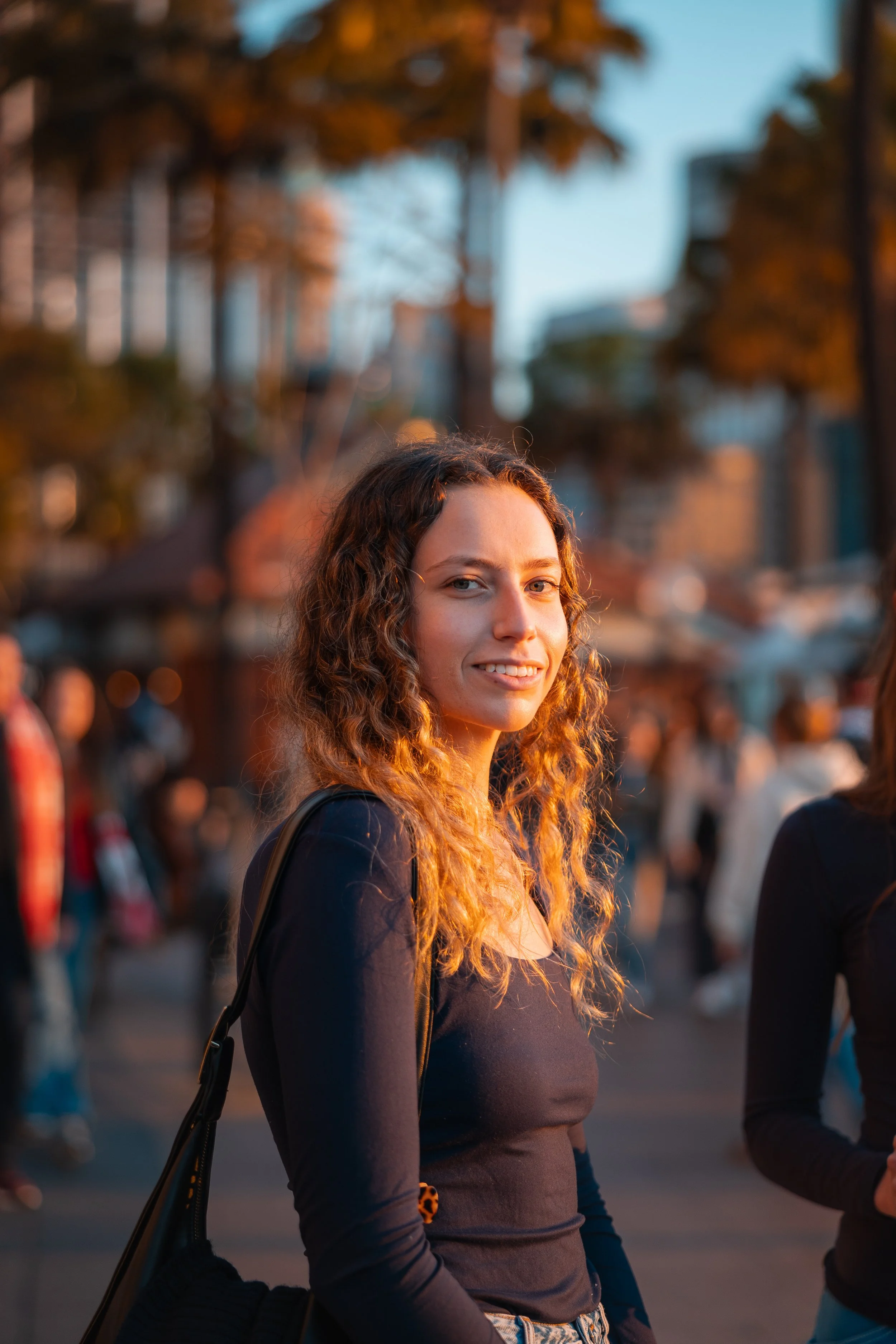 A young woman with curly hair wearing a black shirt and carrying a black bag is smiling outdoors during golden hour, with a crowd and trees in the background.