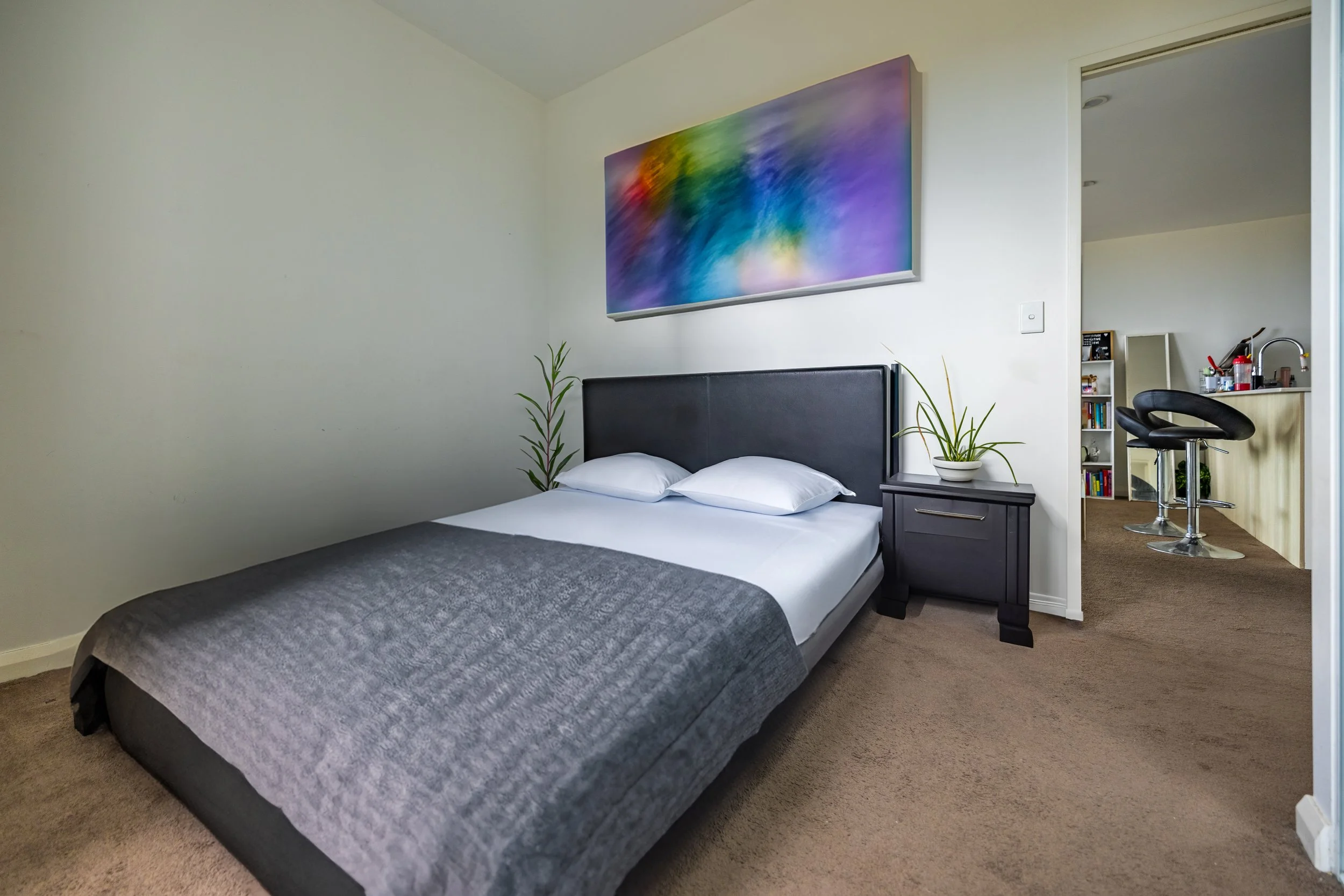 A bedroom with a bed featuring a dark headboard, white pillows, white sheets, and a gray quilt. There are two plants on either side of the bed and a colorful abstract painting above. A door opens to a small kitchen area with bar stools and a bookshel