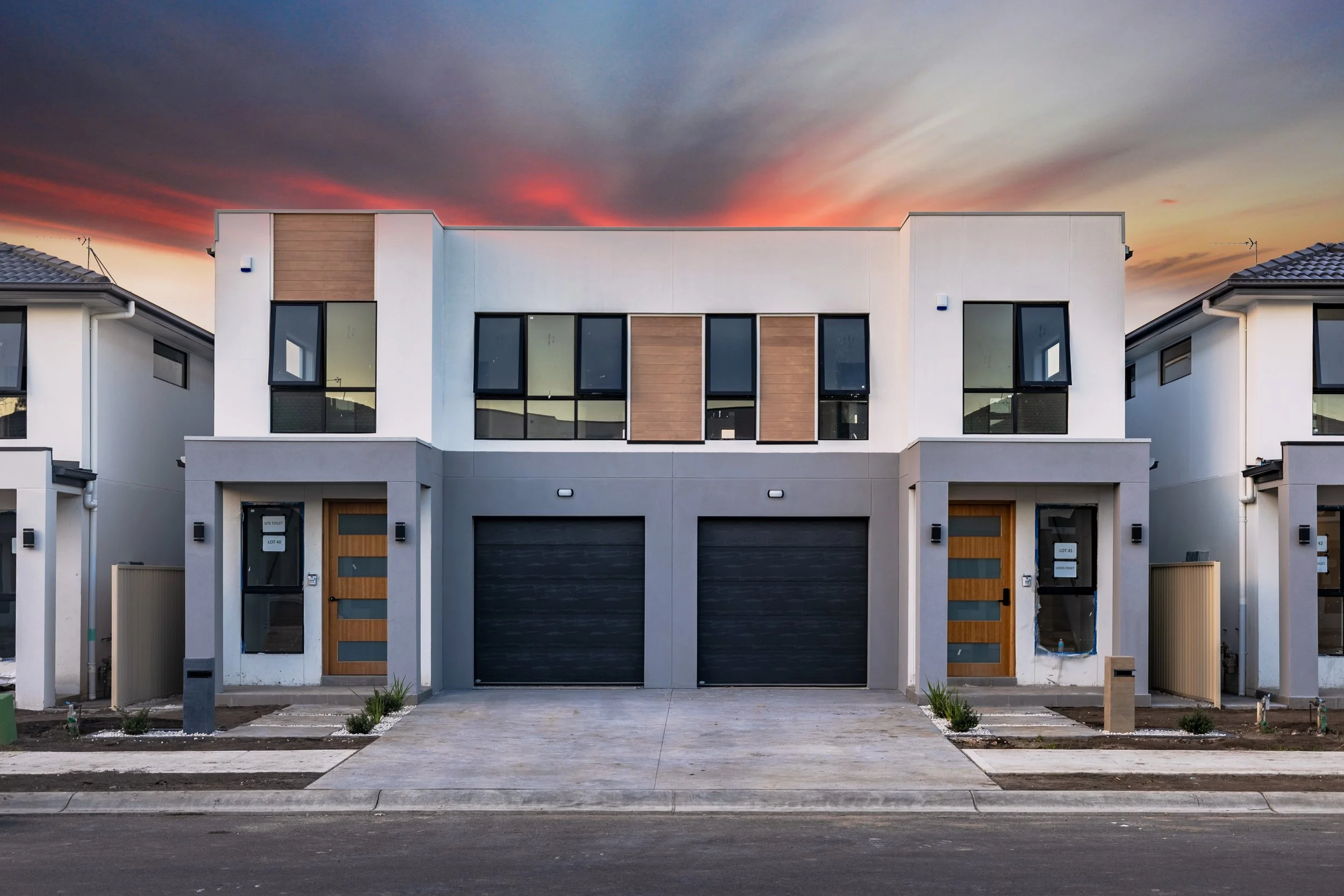Modern two-story residential building with double garage doors and large windows, sunset sky in the background.