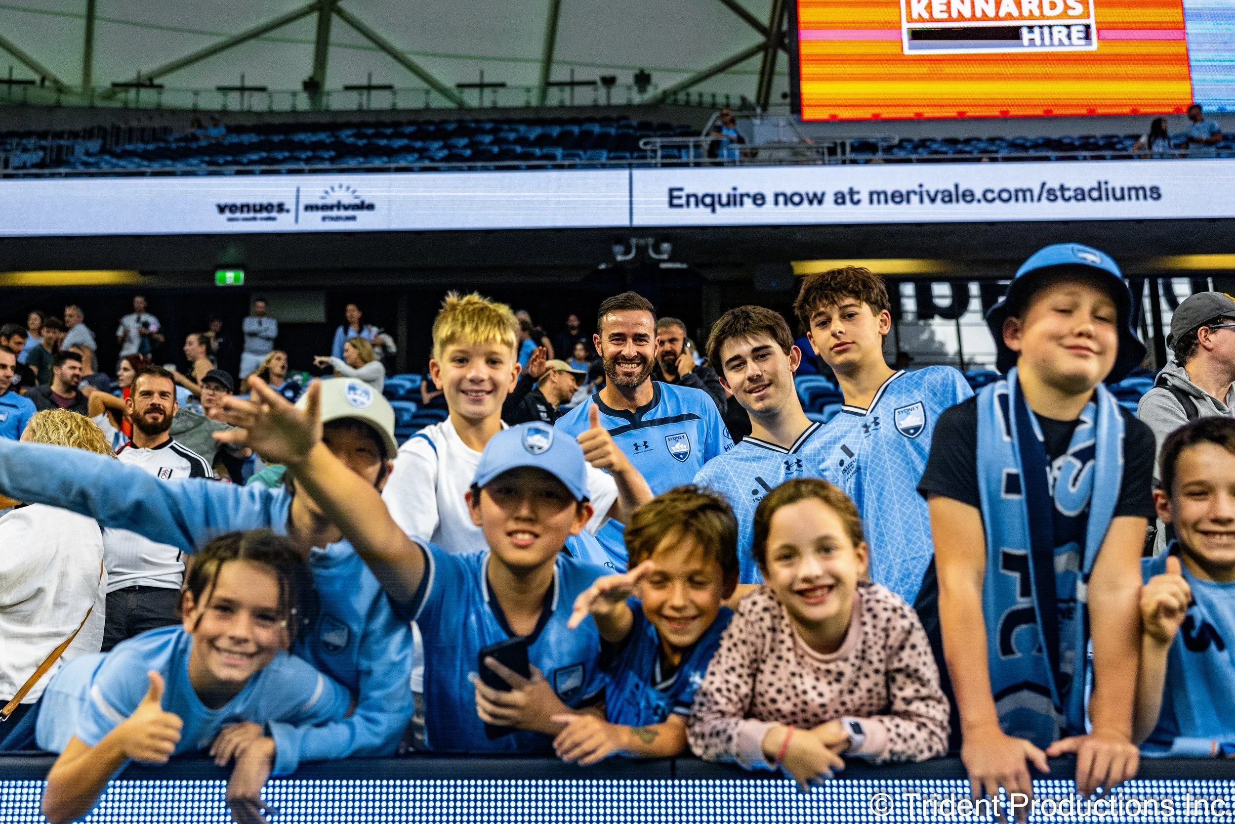 Group of children and a man in a stadium, many wearing Sydney FC jerseys, smiling and posing for a photo.