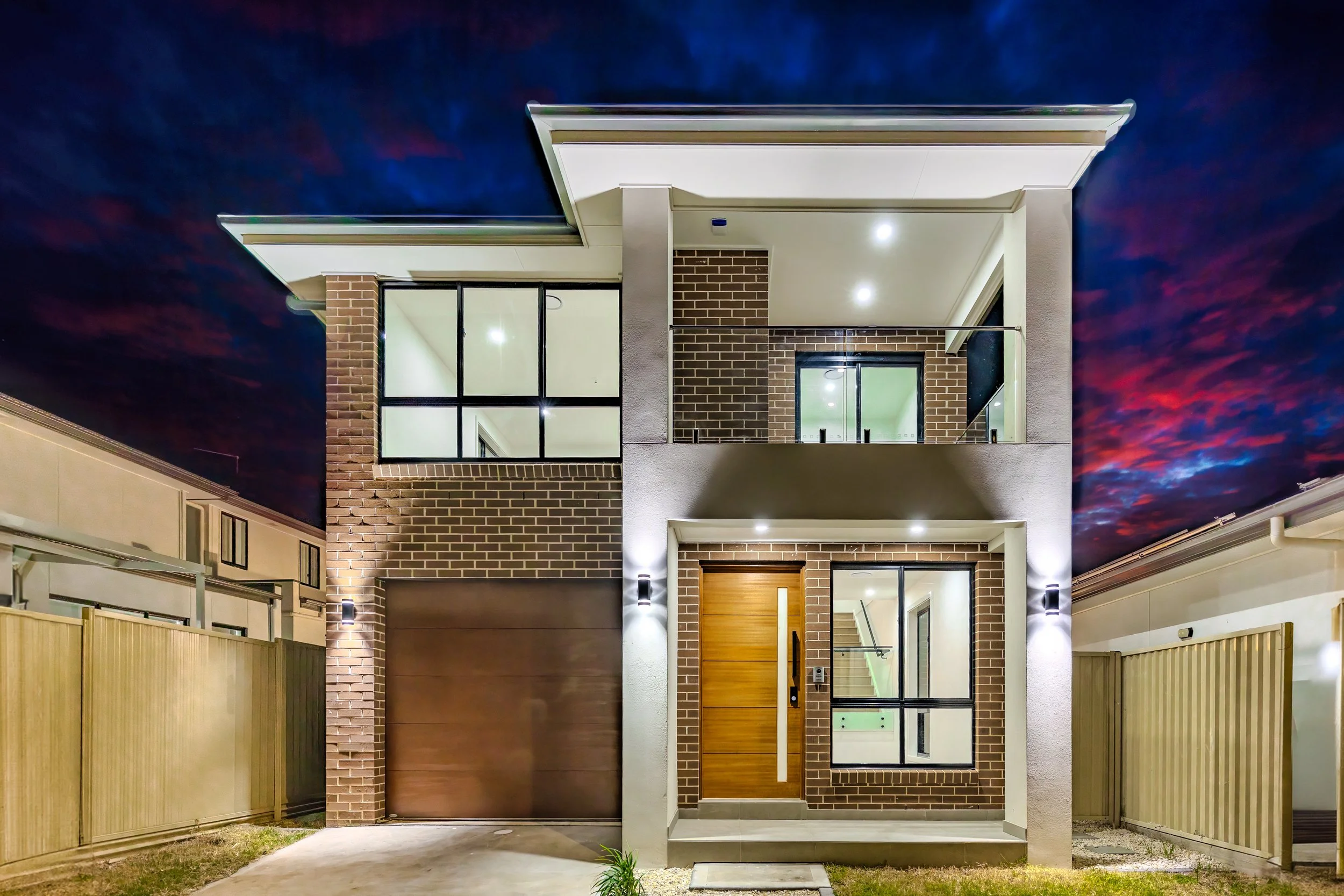 Modern two-story house with brick exterior, large windows, and a wooden front door, illuminated at night against a dark, colorful sky.
