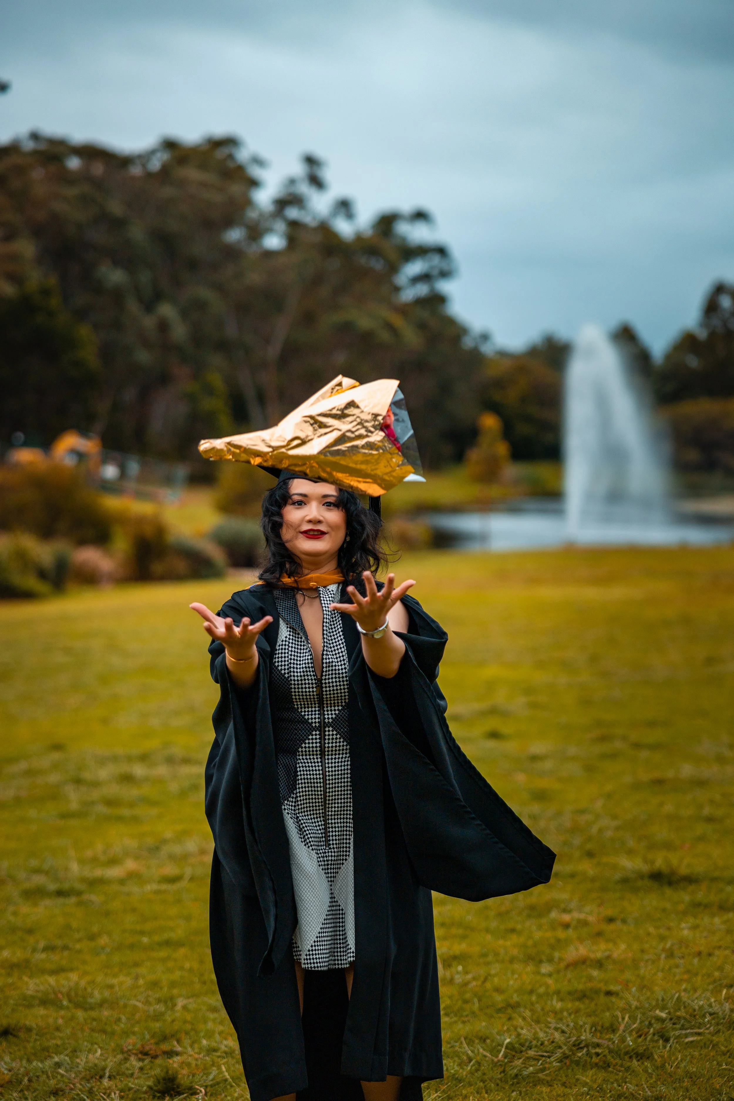 A woman in graduation attire is outdoors with a fountain in the background, wearing a large, gold-colored hat.