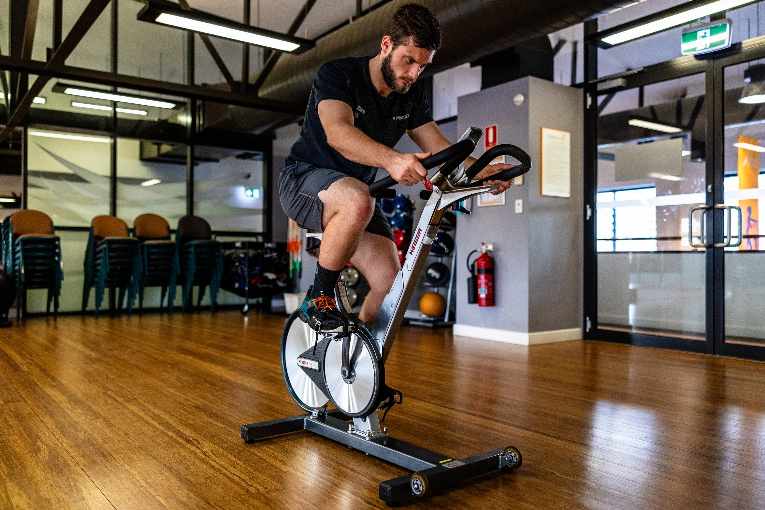 A man is using a stationary exercise bike indoors with wooden floors, chairs in the background, and glass doors to the side.