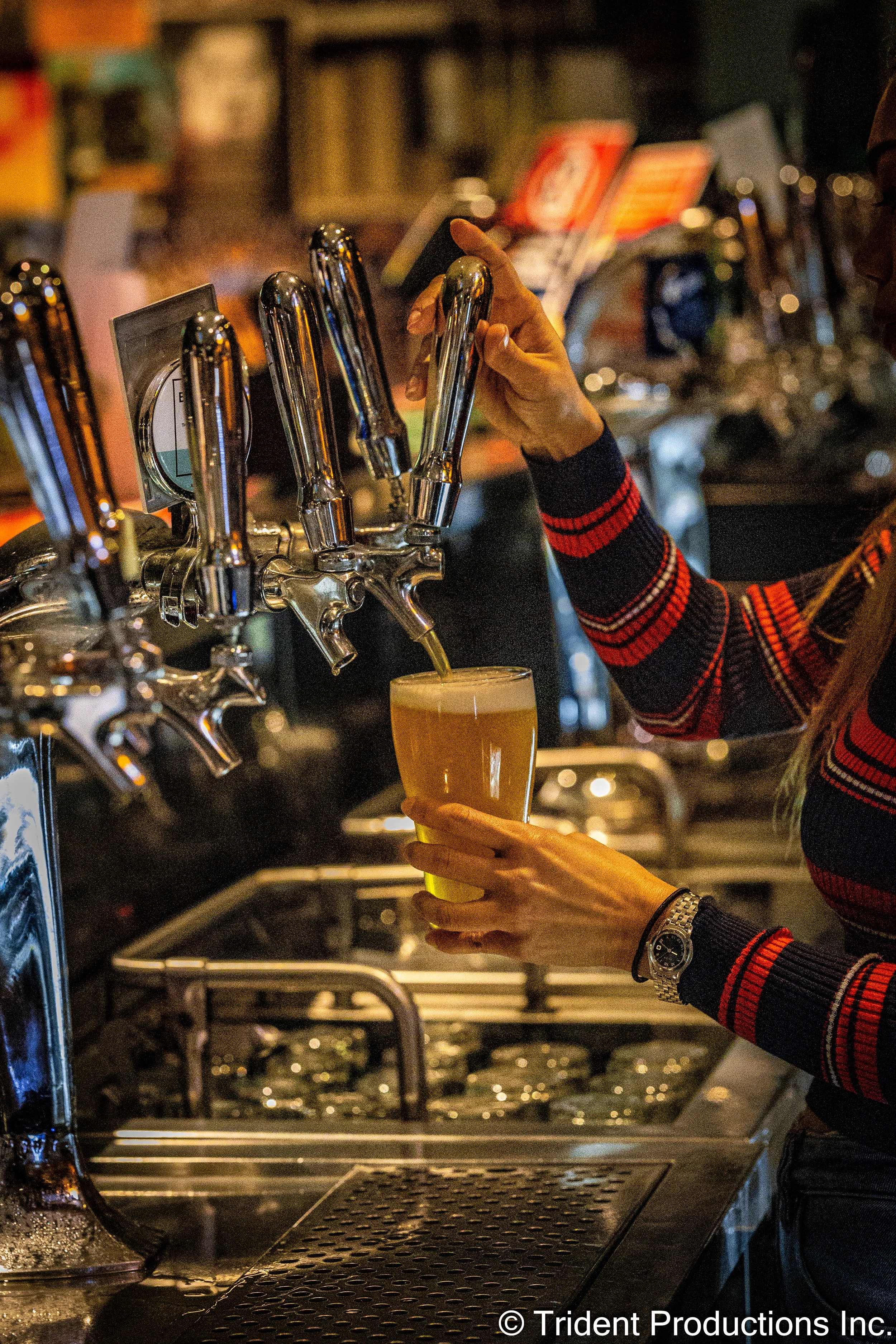A person is pouring beer from a tap into a glass in a bar, wearing a striped sweater and a wristwatch.