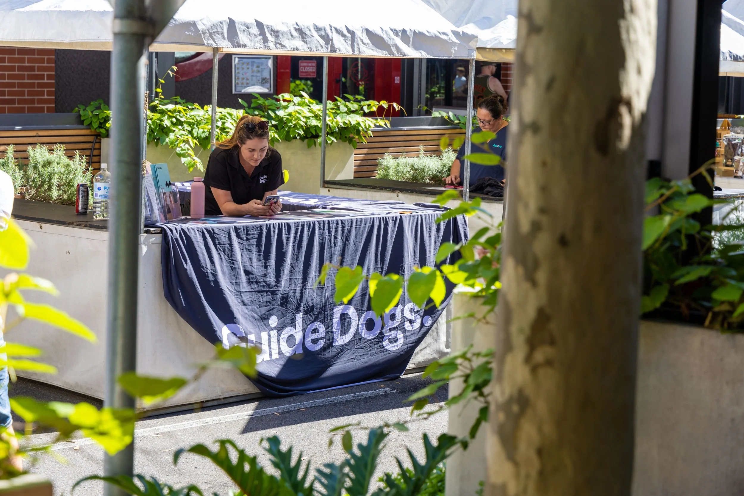 Two women working at a table with a 'Guide Dogs' banner outdoors.