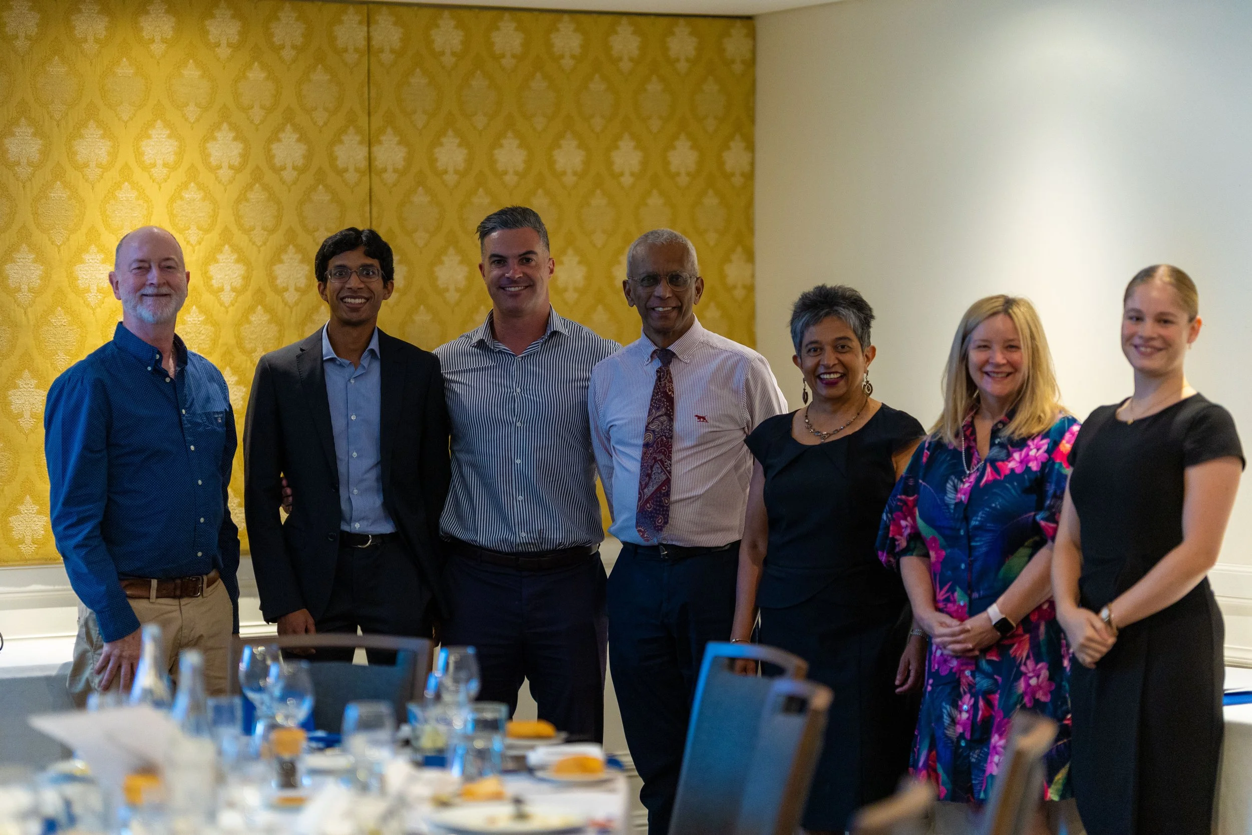 A group of eight professionally dressed people standing together at a formal event in a banquet hall with yellow patterned walls.