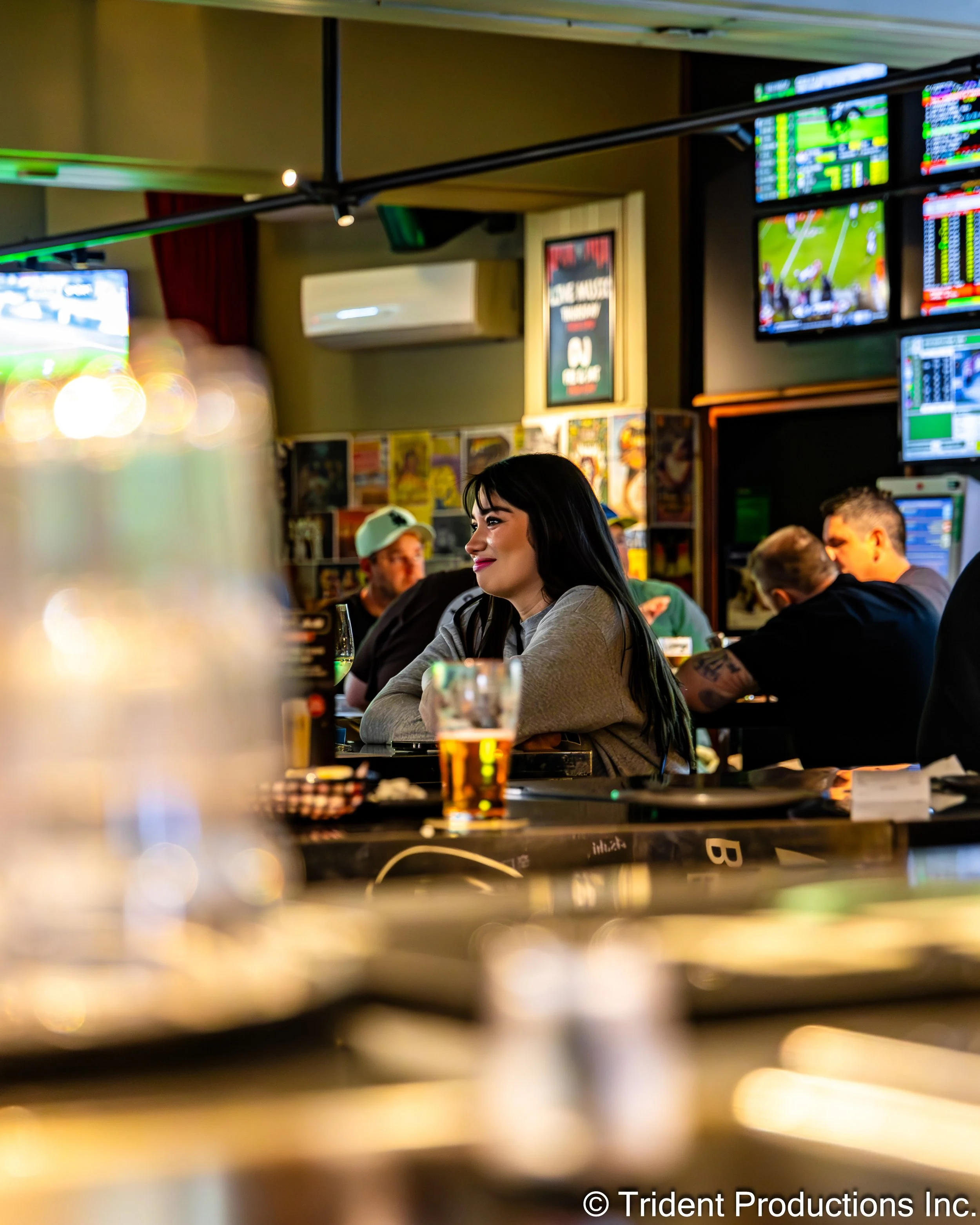 A woman with long dark hair sitting at a bar in a sports bar, smiling and watching a game on TVs mounted on the wall. There are other patrons, drinks, and sports-related decor in the background.
