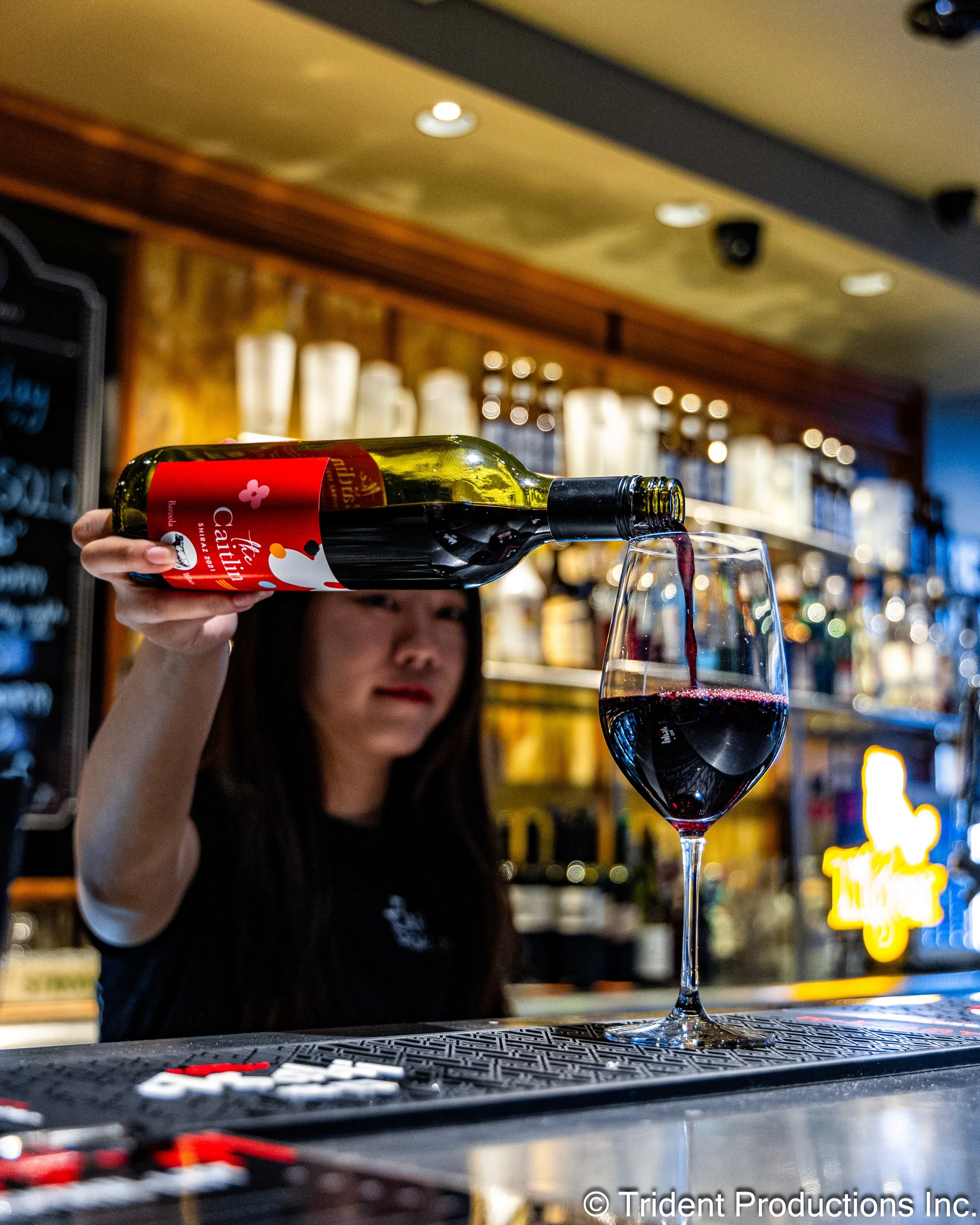 A woman is pouring red wine from a bottle into a wine glass at a bar. The background shows shelves with bottles and bar decorations.