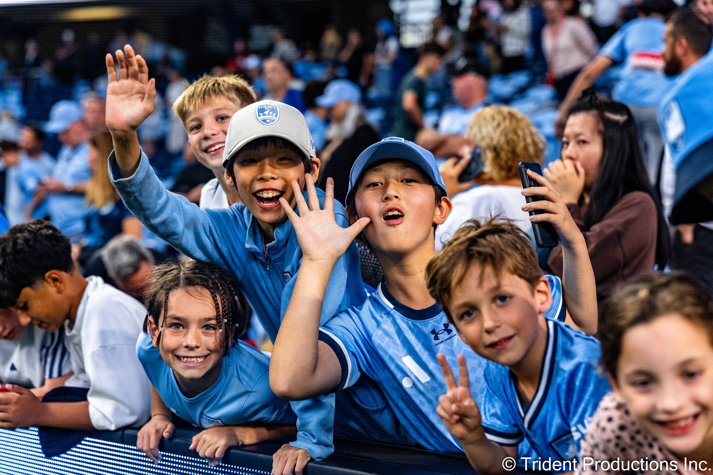 A group of children and adults at a sporting event, wearing blue jerseys and hats, smiling and waving at the camera.