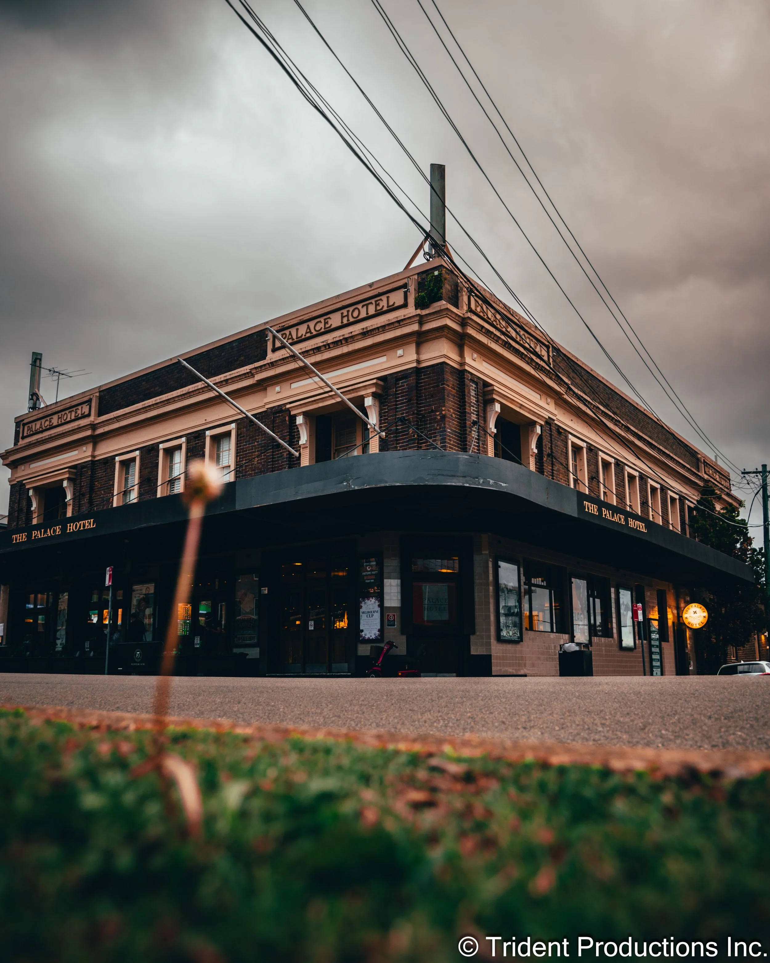 Photo of a brick hotel building with a sign that reads "The Palace Hotel." The building has multiple windows and a corner entrance, with an overcast sky and power lines overhead.