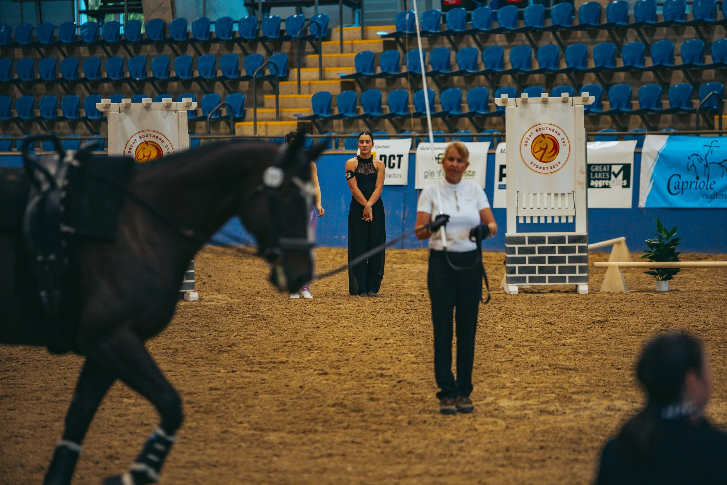 An indoor equestrian arena with blue seating, showing a woman riding a dark horse, and a woman with a clipboard in the foreground.