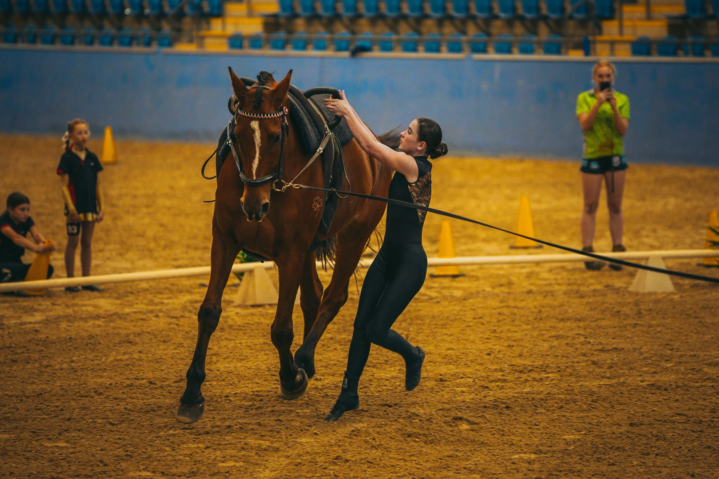 A woman in a black outfit tending to a brown horse in an indoor riding arena, with several children and a woman taking photos in the background.