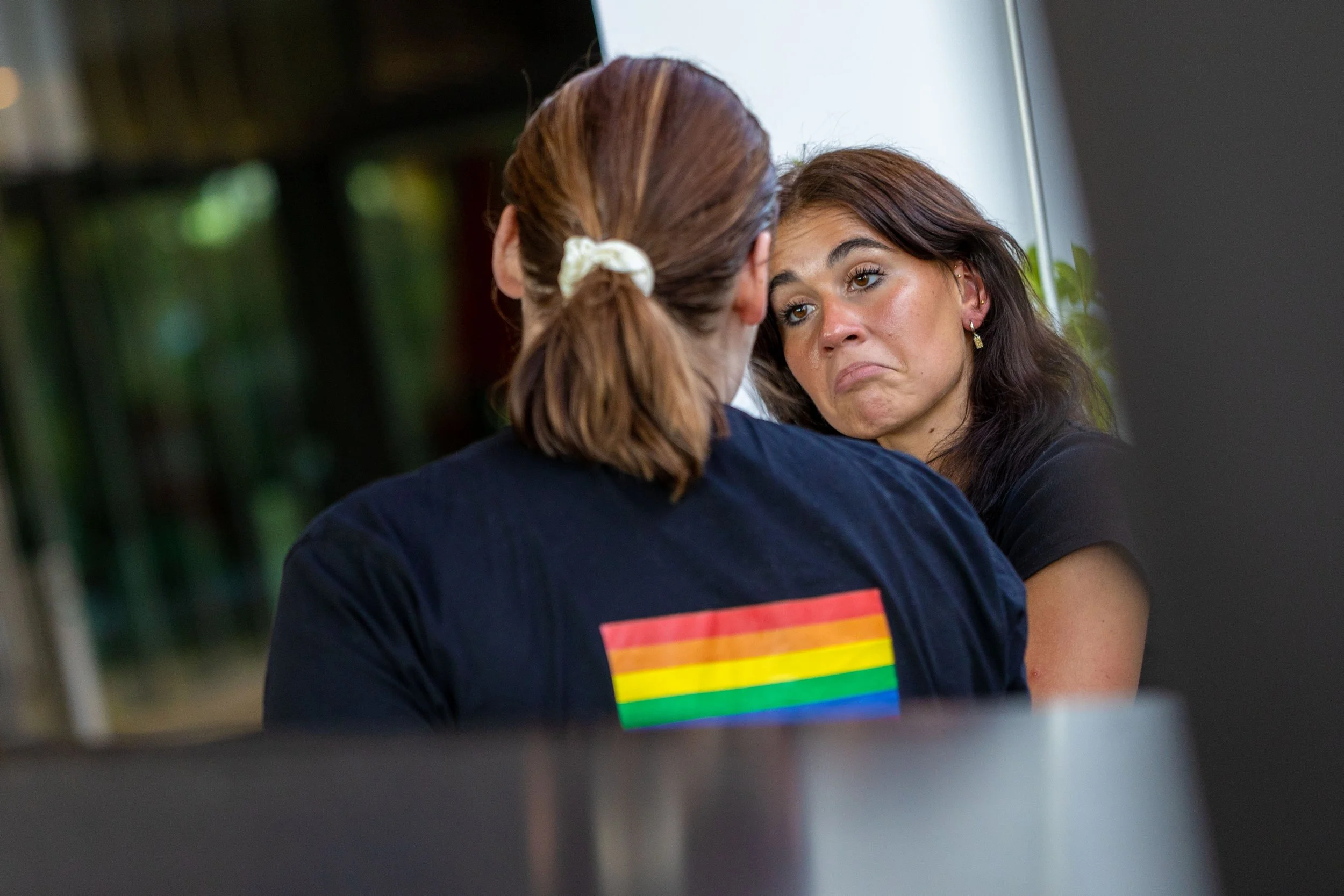 A woman with brown hair and a black shirt looks upset as she is comforted by another woman with brown hair tied in a ponytail, wearing a black shirt with a rainbow pride flag on the back.
